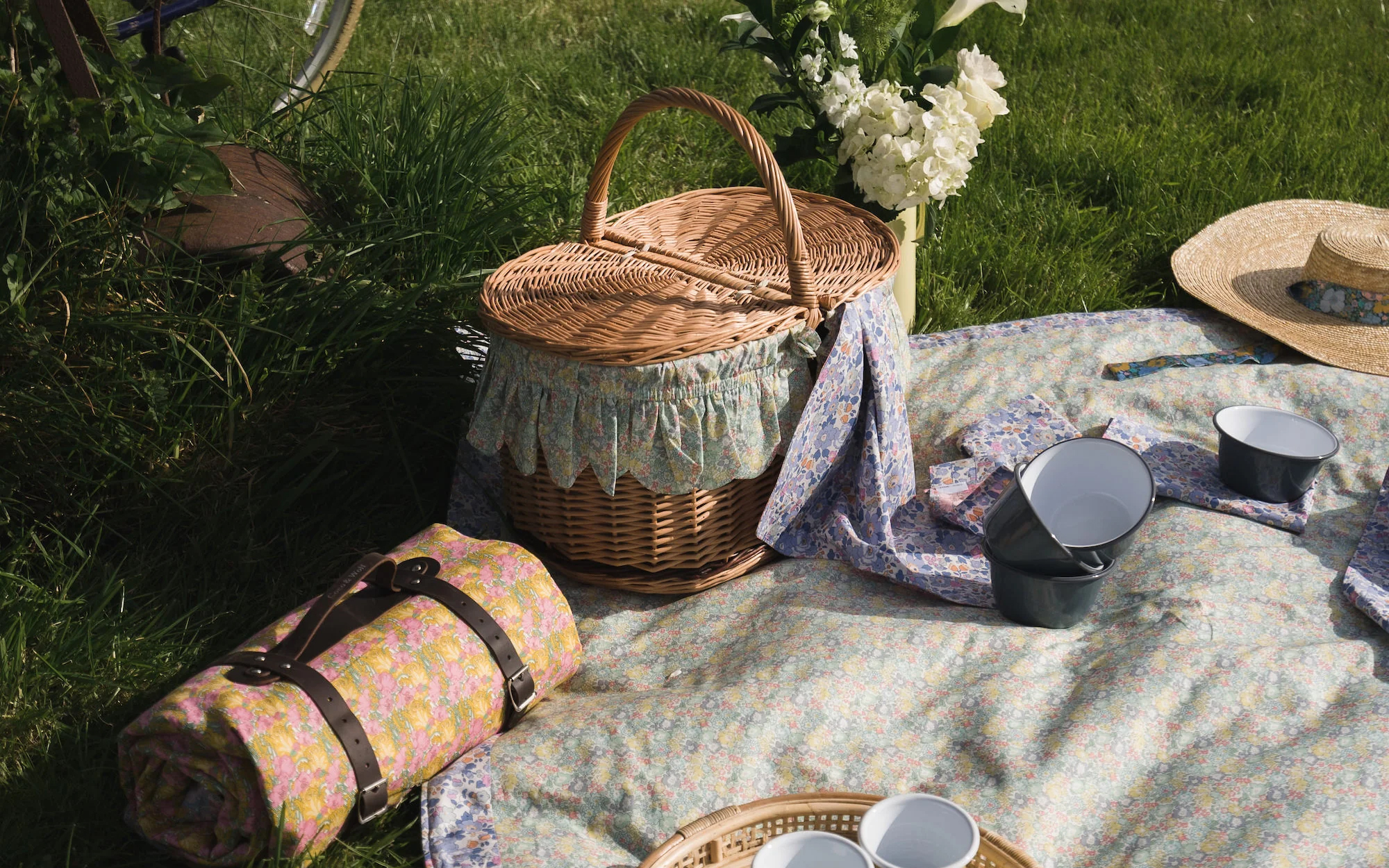a picnic basket with a floral lining on a pale floral picnic blanket, a rolled up pink blanket and a vase of white flowers on the grass