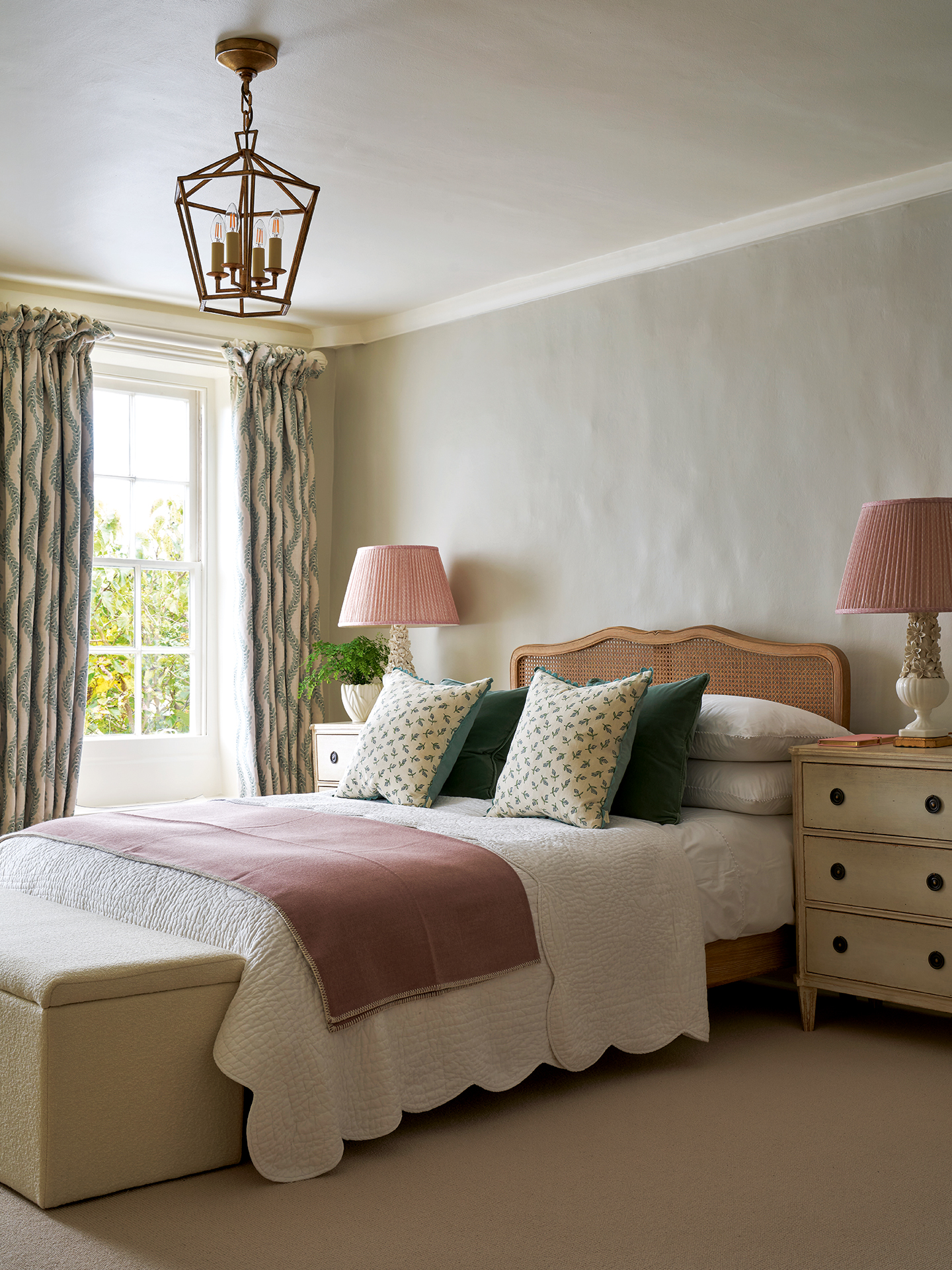 A light blue-grey bedroom with a rattan double bed, decorated with a quilted white bedcover, pink throw and dainty floral cushions, large bedside tables with drawers, topped with table lamps with pleated pink shades, a storage bench and floral curtains 