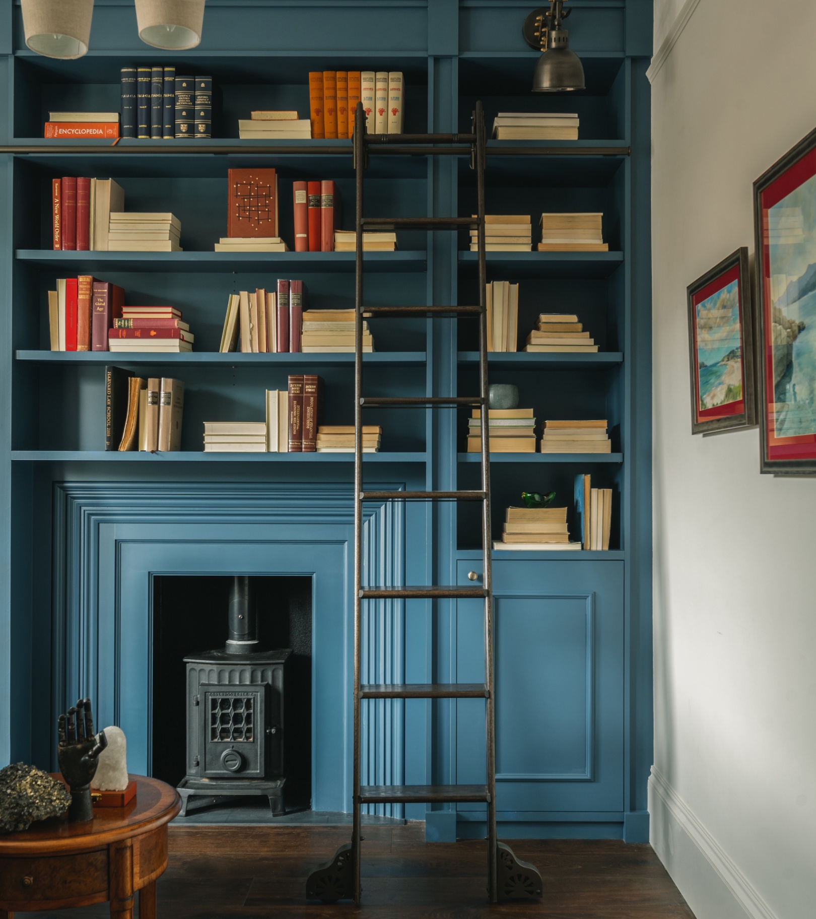 a living room corner with a floor to ceiling blue painted bookcase full of books and a library ladder infront of it with a built in log burner