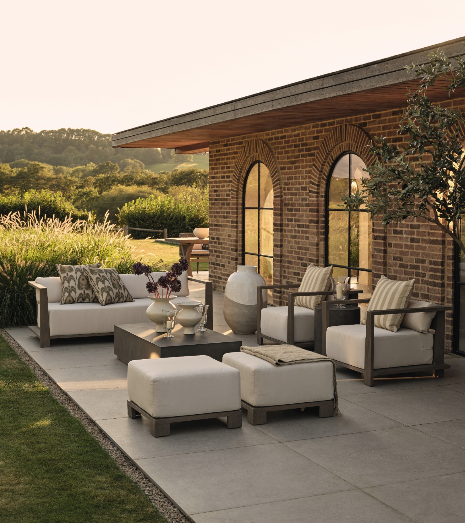 a patio with a set of luxury outdoor furniture with cream seat pads and white and green stripe cushions against a red brick house surrounded by trees and a lawn