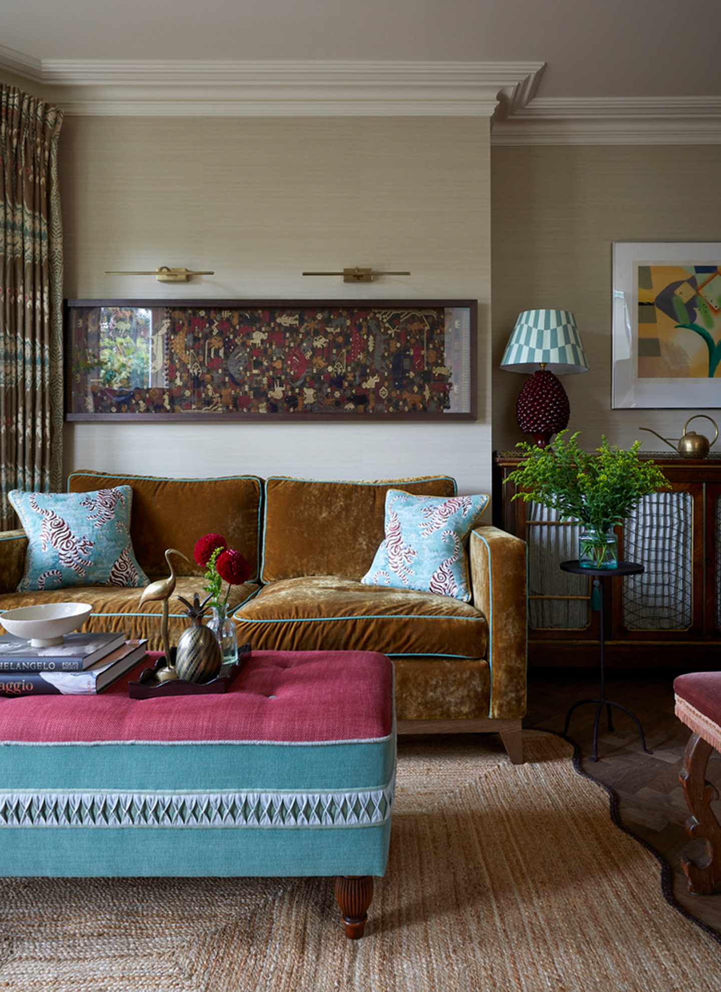 A sophisticated living room with neutral walls, a mustard yellow velvet sofa, light blue patterned cushions, a bright pink and turquoise upholstered ottoman, and a jute rug underfoot