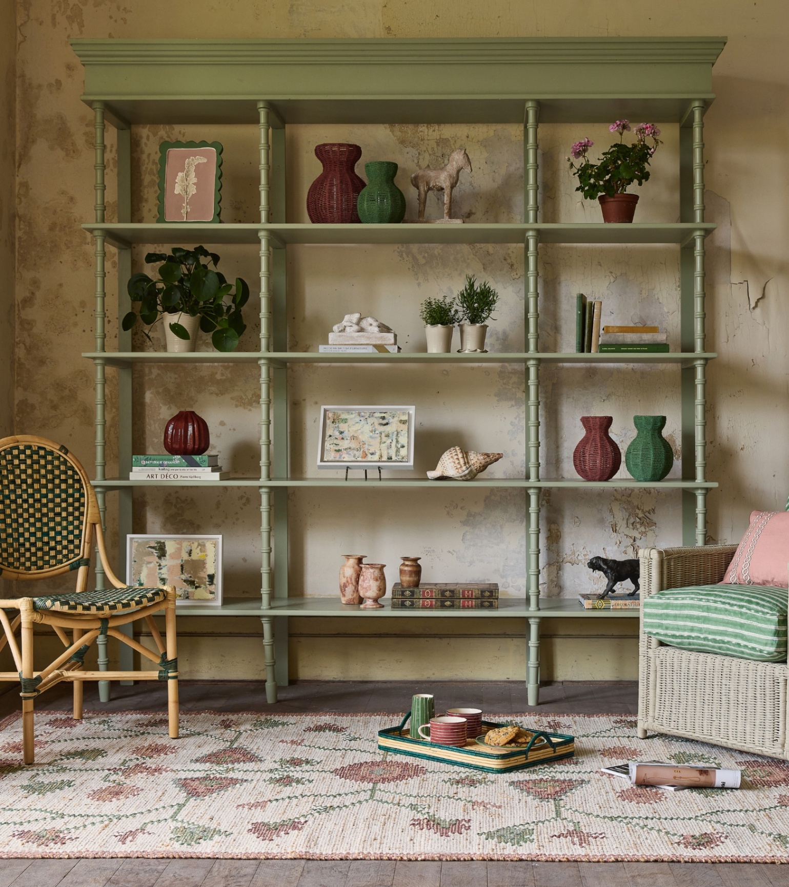 a large sage green painted shelving unit filled with items like wicker vases, shells, ornaments and books with a red green and white patterned rug on the floor and a bistro chair to the left and a grey rattan chair to the right with a green stripe cushion