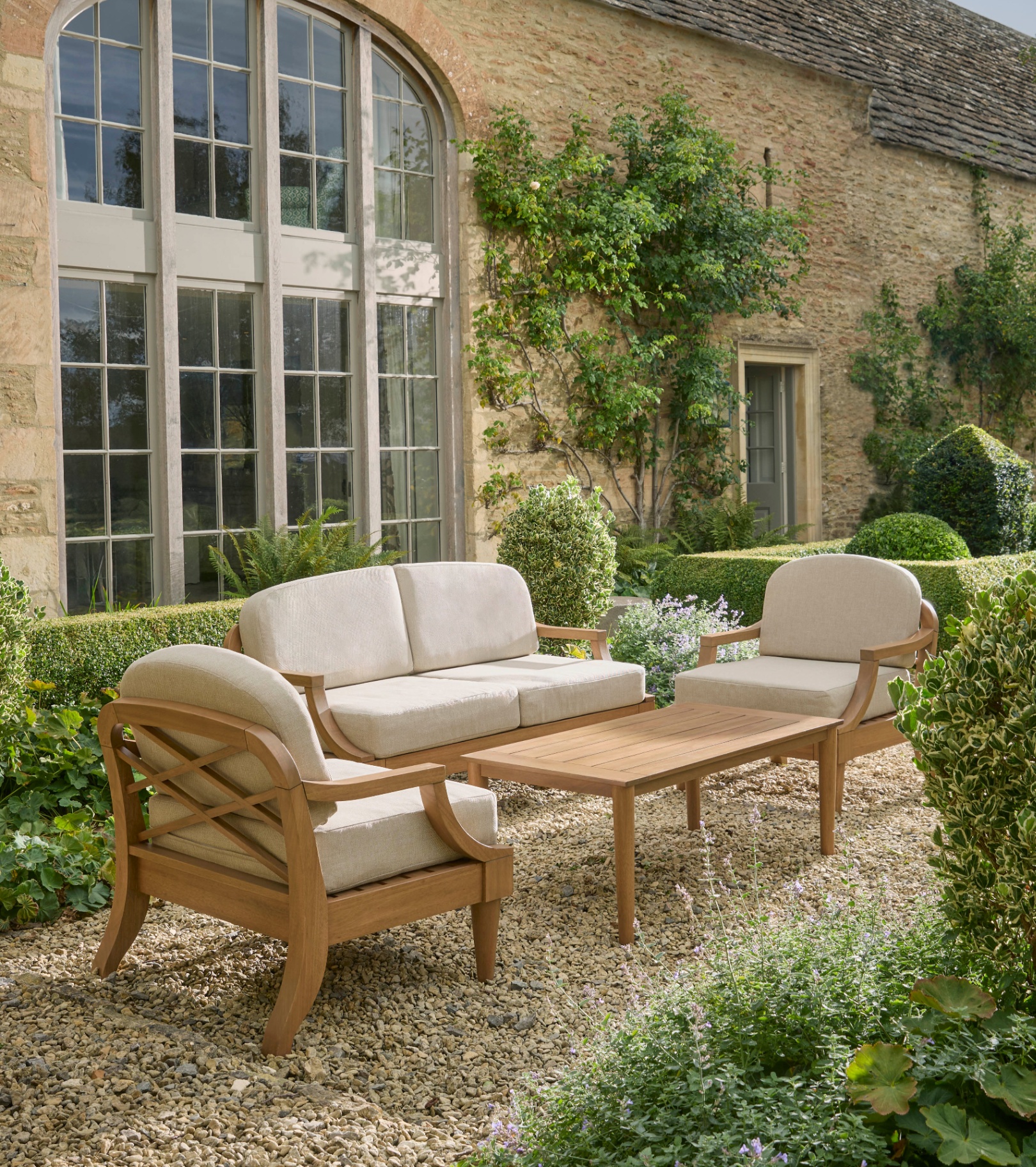 An outdoor garden furniture set with wooden frame and beige seat pads on a country house patio surrounded by greenery