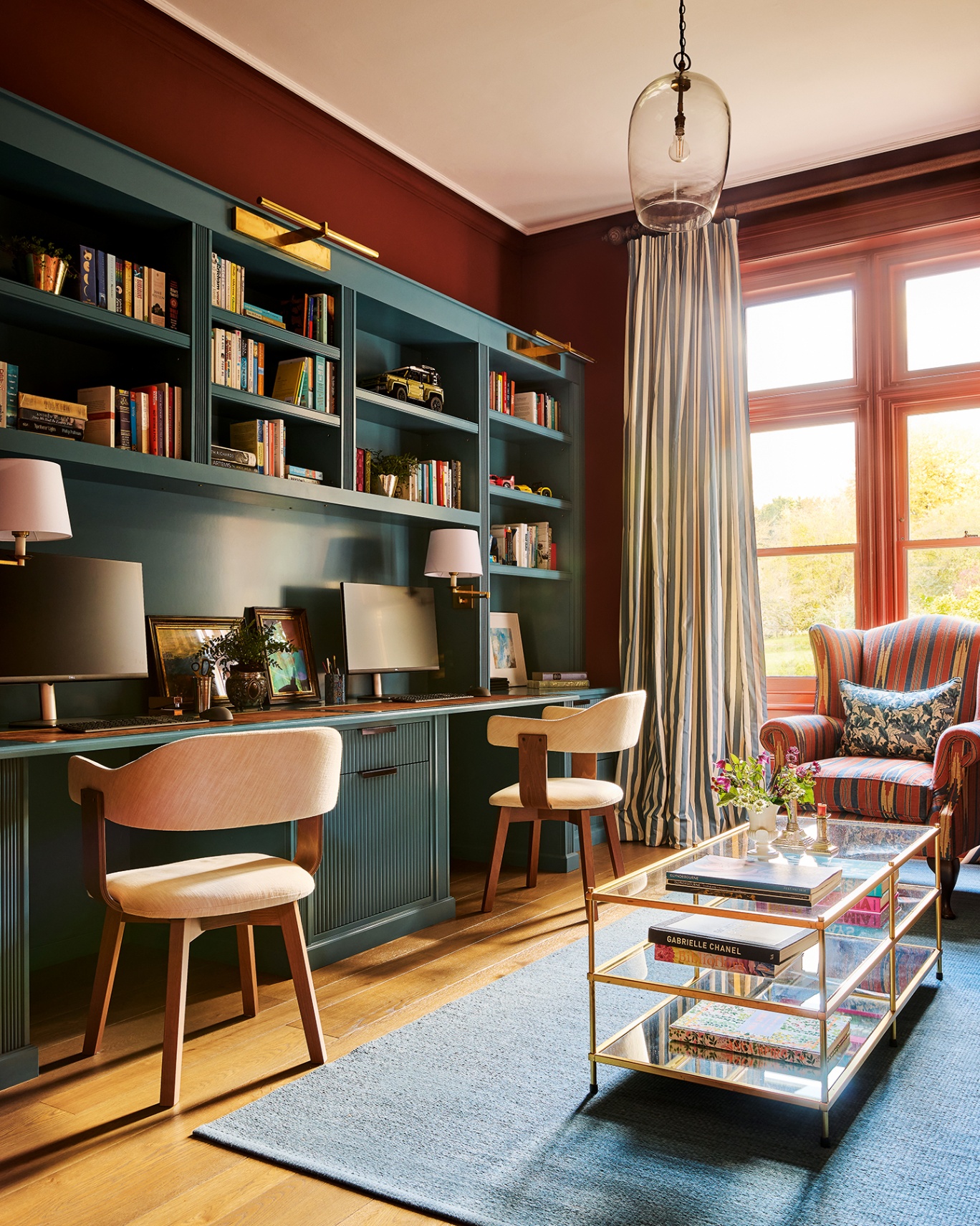 A home office with dark red walls and a built-in blue wall unit. It is filled with books and on the desk level sit two computers. There are two desk chairs. In the foreground is a blue rug and gold and glass coffee table, and behind is a striped armchair
