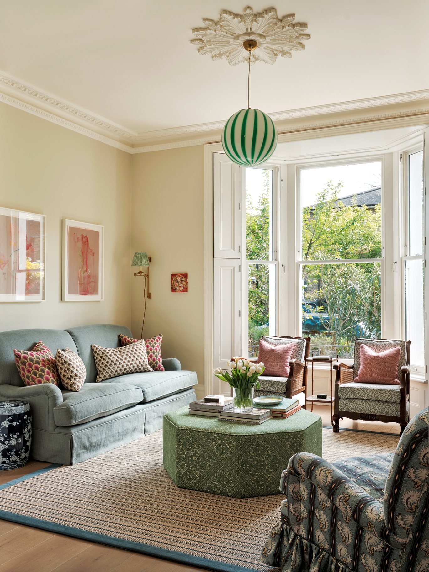 a living room with an octagonal green ottoman surrounded by three armchairs and a blue sofa, all sat on a jute rug with a blue border. A large bay window looks out onto trees and a striped green and white globe pendant hangs from an ornate ceiling rose