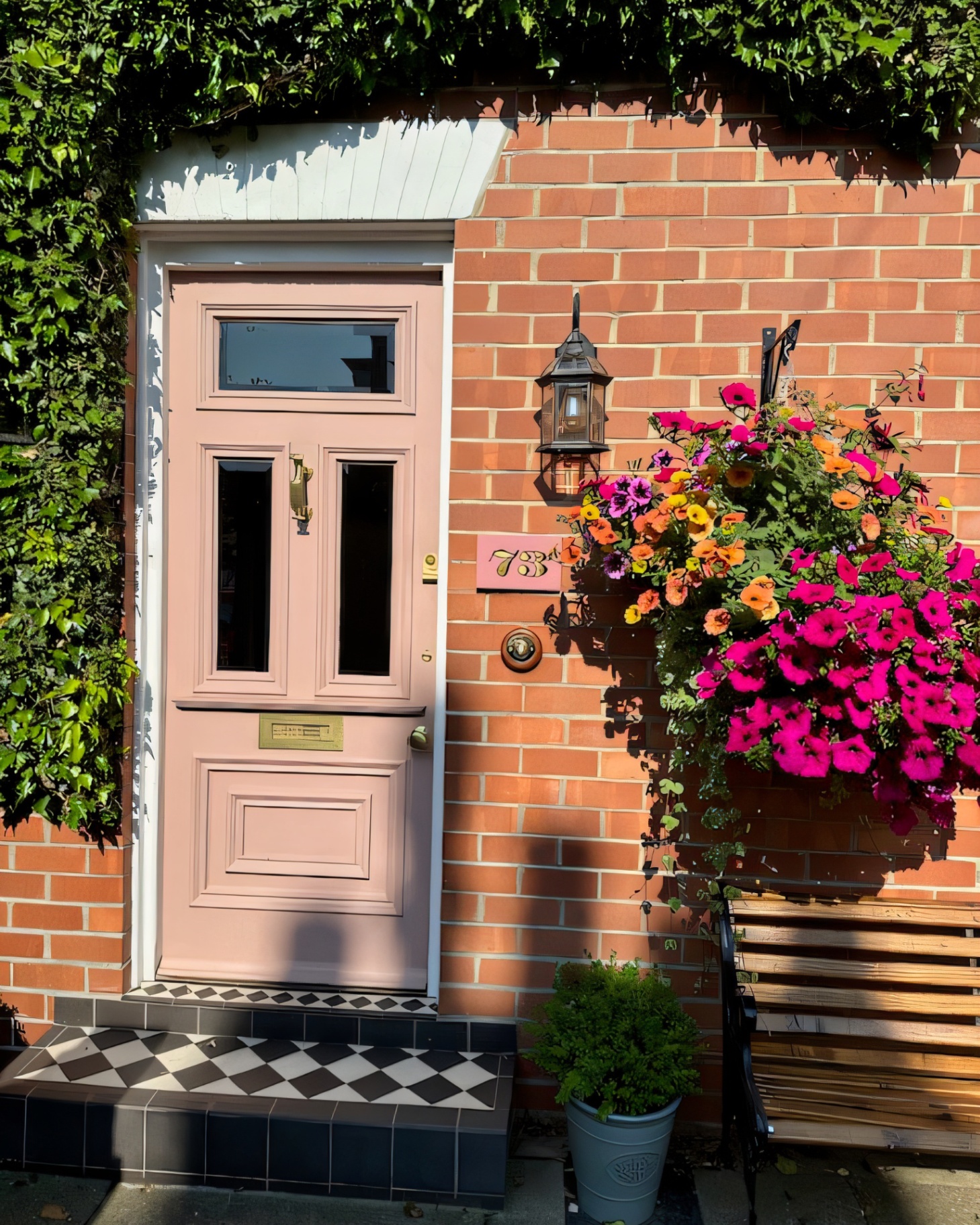 a red brick house with a pale pink front door, a black and white checkerboard tiled step, a pink door number tile and a hanging basket with colourful flowers