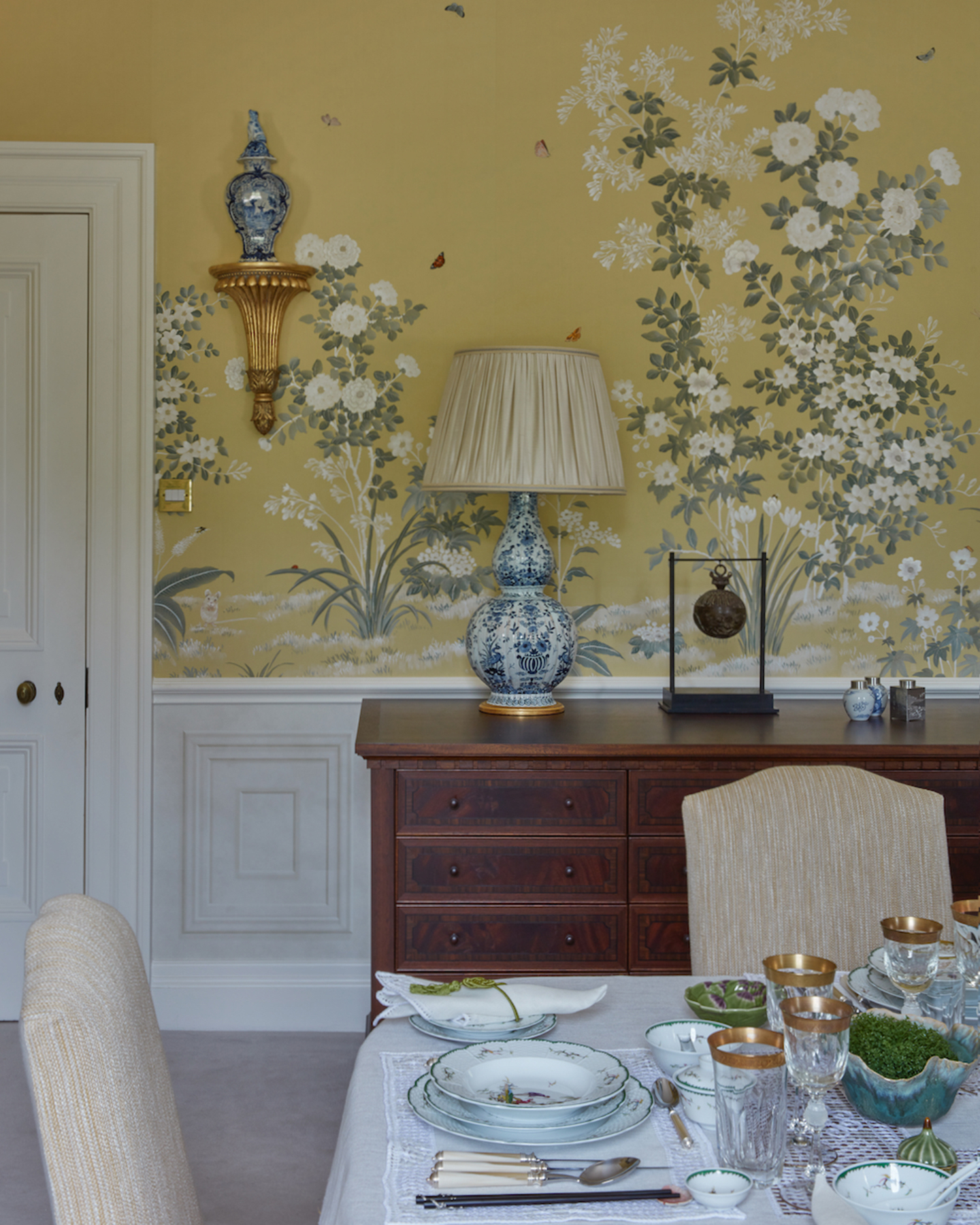 a dining room with a yellow wall covered in a handpainted flower mural, an antique dark wood side board with a blue and white floral lamp and a dining table with wine glasses and decorative plates
