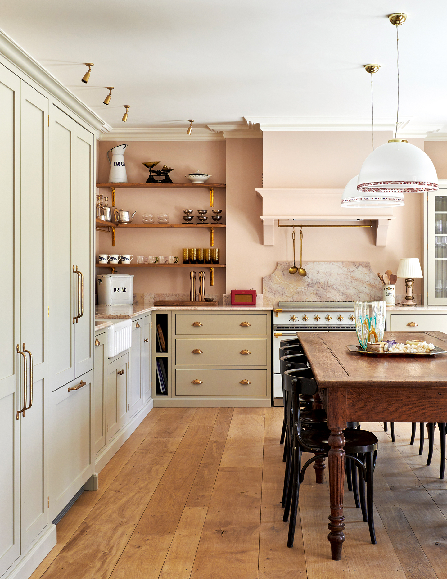 A pink kitchen with wooden floorboards, neutral cabinetry and a cooker with a marble backsplash. In the foreground, a large wooden dining table is paired with black cafe chairs and two large glass dome pendant lights