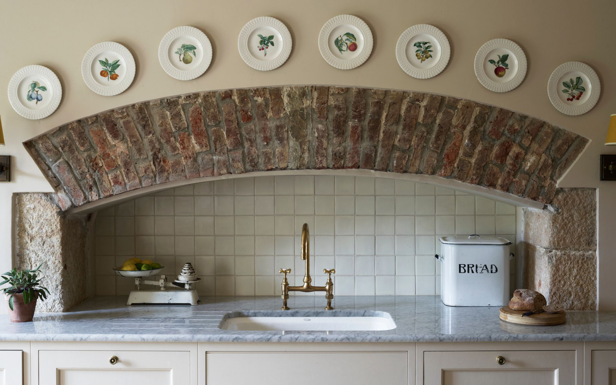 a country style kitchen sink area, with gold taps and an exposed brick arch alcove with decortive plates lined above it