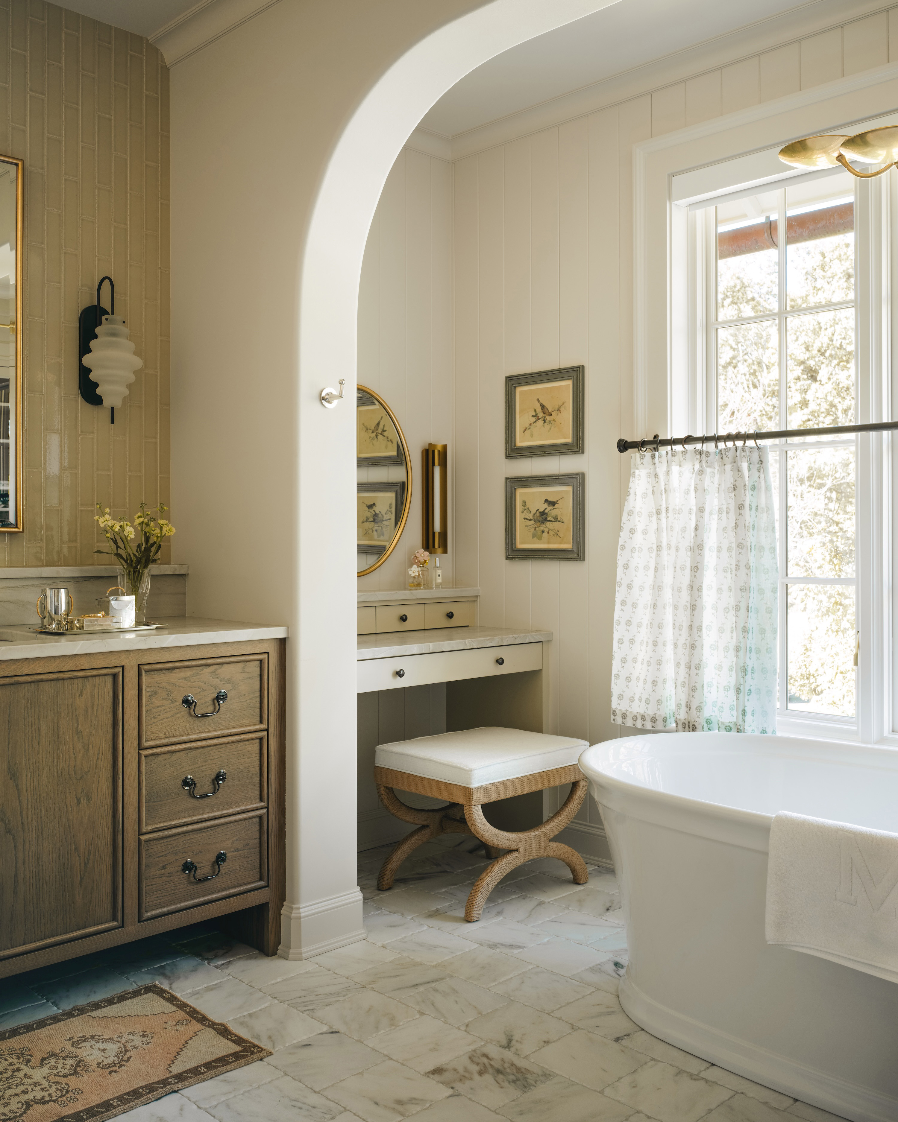 a neutral bathroom with a dressing room area at the back left next to a large window, a dark wooden vanity with a mirror and wall lights in the foreground and a large white bath to the right
