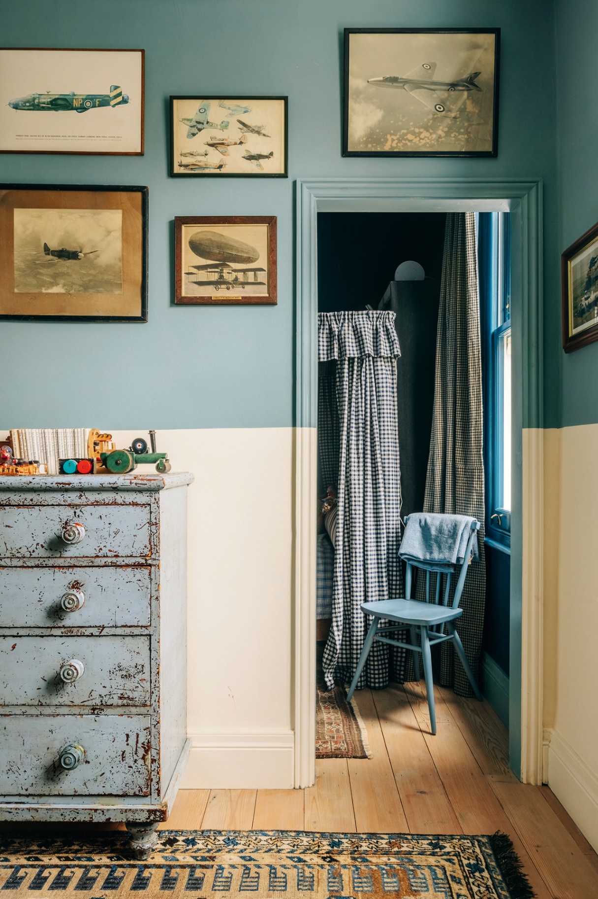 in the foreground a vintage chest of drawers is placed against a blue and white half-and-half painted wall decorated with vintage pictures of aircrafts. Through the door a glimpse of a bedroom can be seen with a blue chair and gingham bed canopy