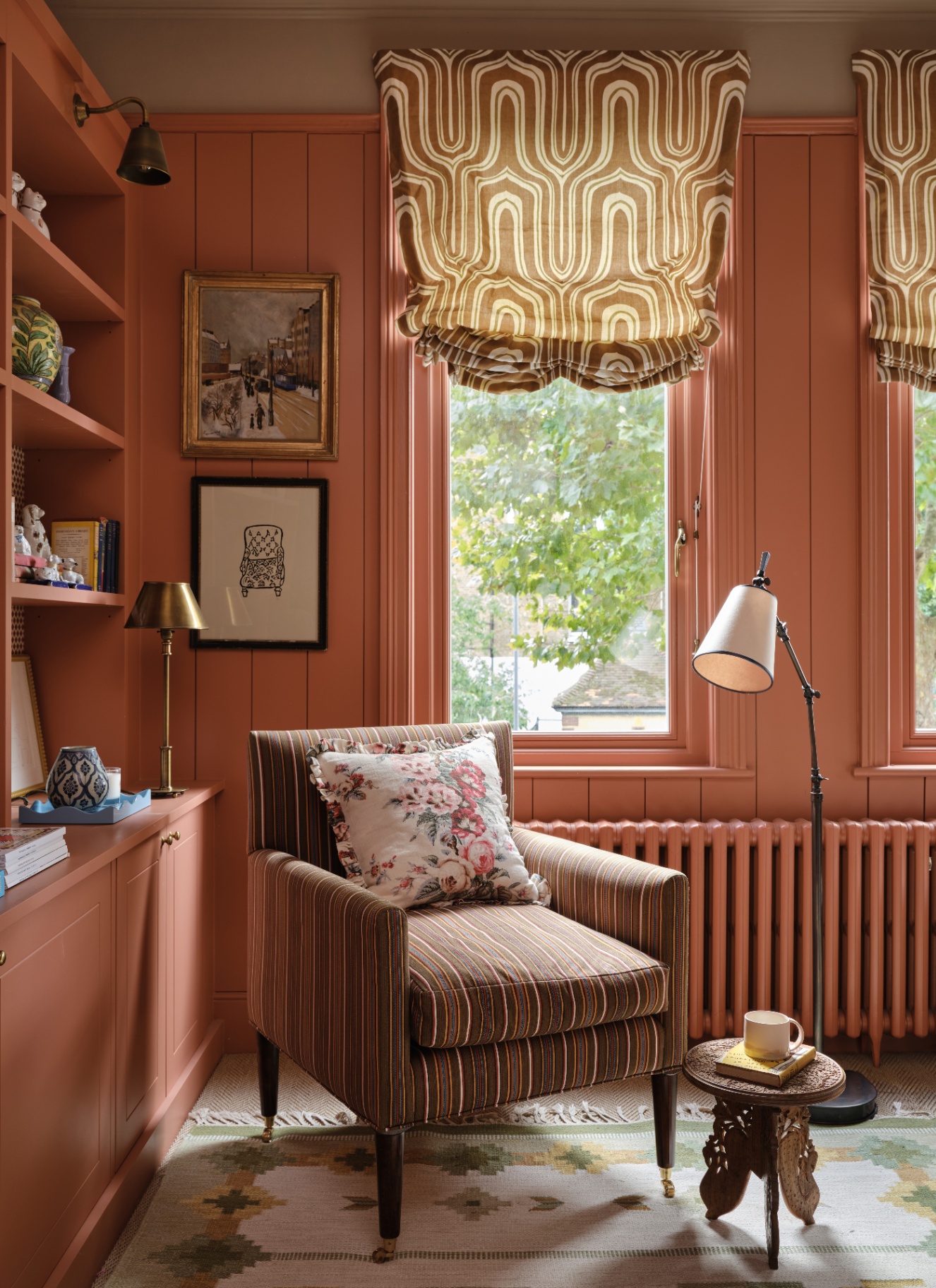 A warm, terracotta-hued reading nook with matching wall panelling and radiator, a striped armchair, floral cushion, Roman blinds, and a brass task lamp
