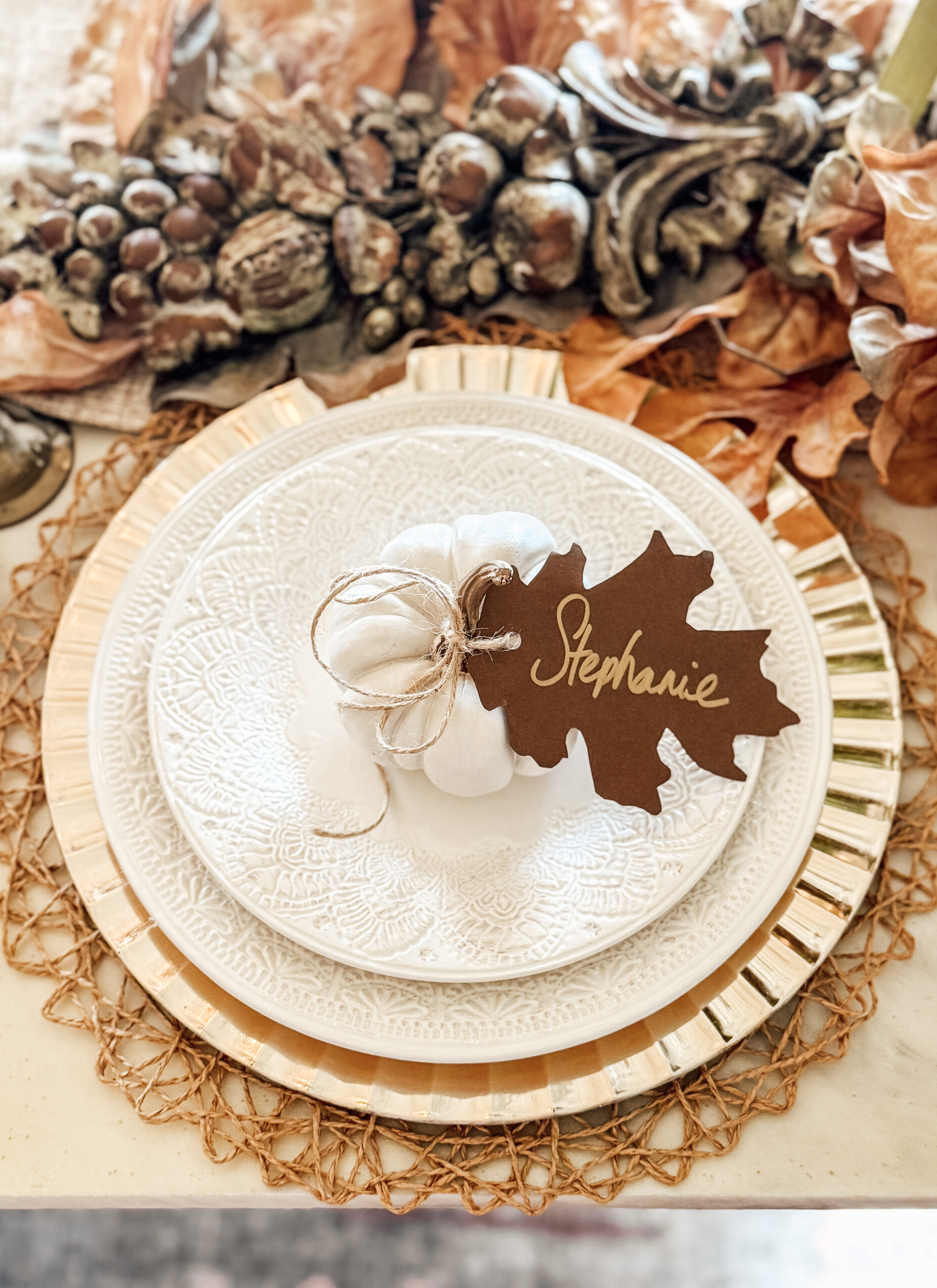 close up of an autumn table with a stack of white lace patterned plates on a rattan charger plate with a small white pumpkin on top with a brown oak leaf shaped name setting with the name stephanie tied to it and a garland of pinecones behind it