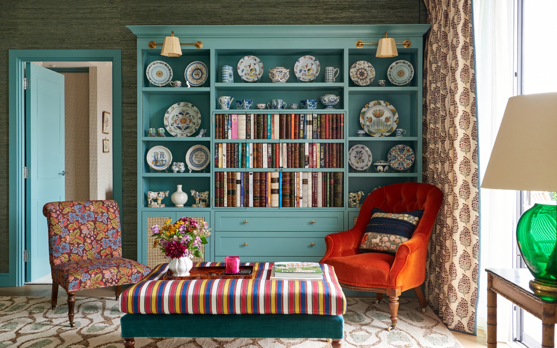 A colourful sitting room featuring a turquoise painted bookcase styled with books and decorative china, flanked by a rich orange velvet armchair and a floral accent chair around a striped ottoman