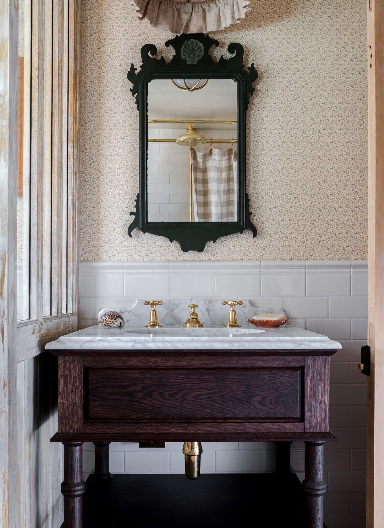 a bathroom with a dark wood antique washstand, gold tap, a large black ornate mirror and cream botanical wallpaper