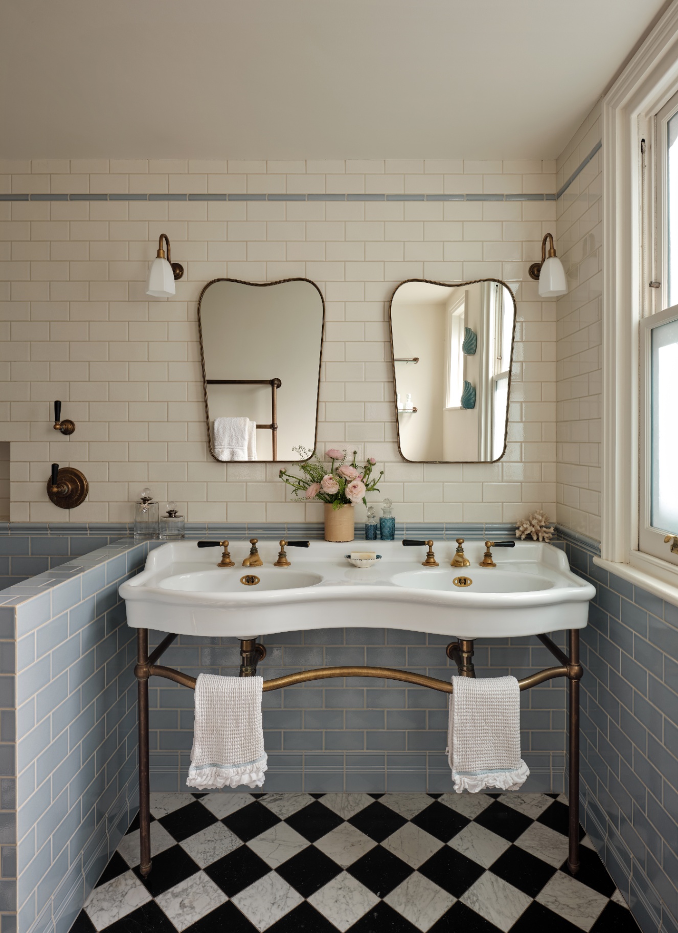 A double console sink with brass fixtures sits against off-white subway tile, topped by twin curved mirrors and sconces. Light blue tiles wrap the lower walls above black-and-white checkered floor tiles, with pink flowers set between the basins