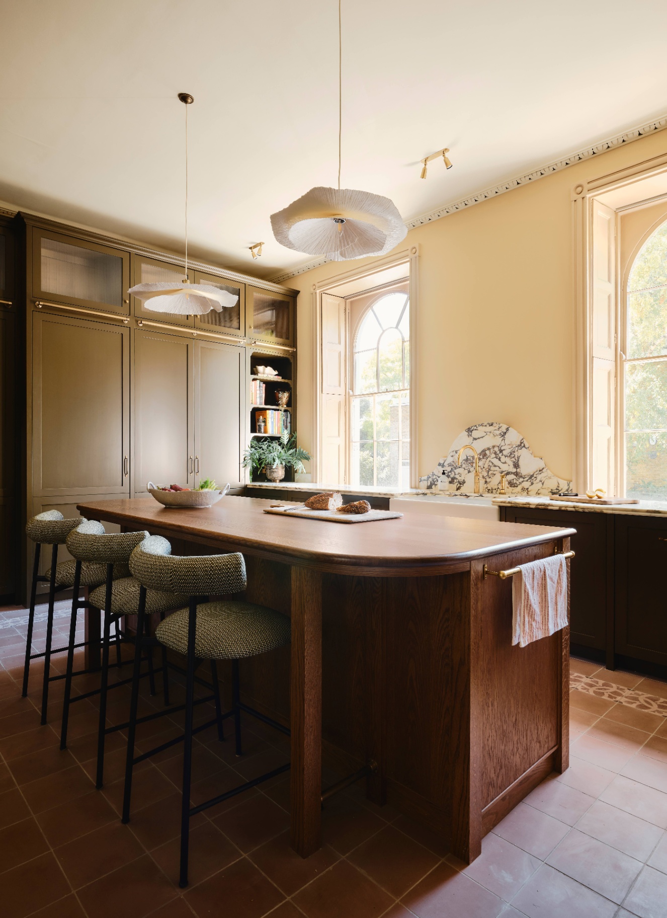 a kitchen with butter yellow walls, a large wooden kitchen island with three upholstered bar stools, and two petal shaped pendant lights