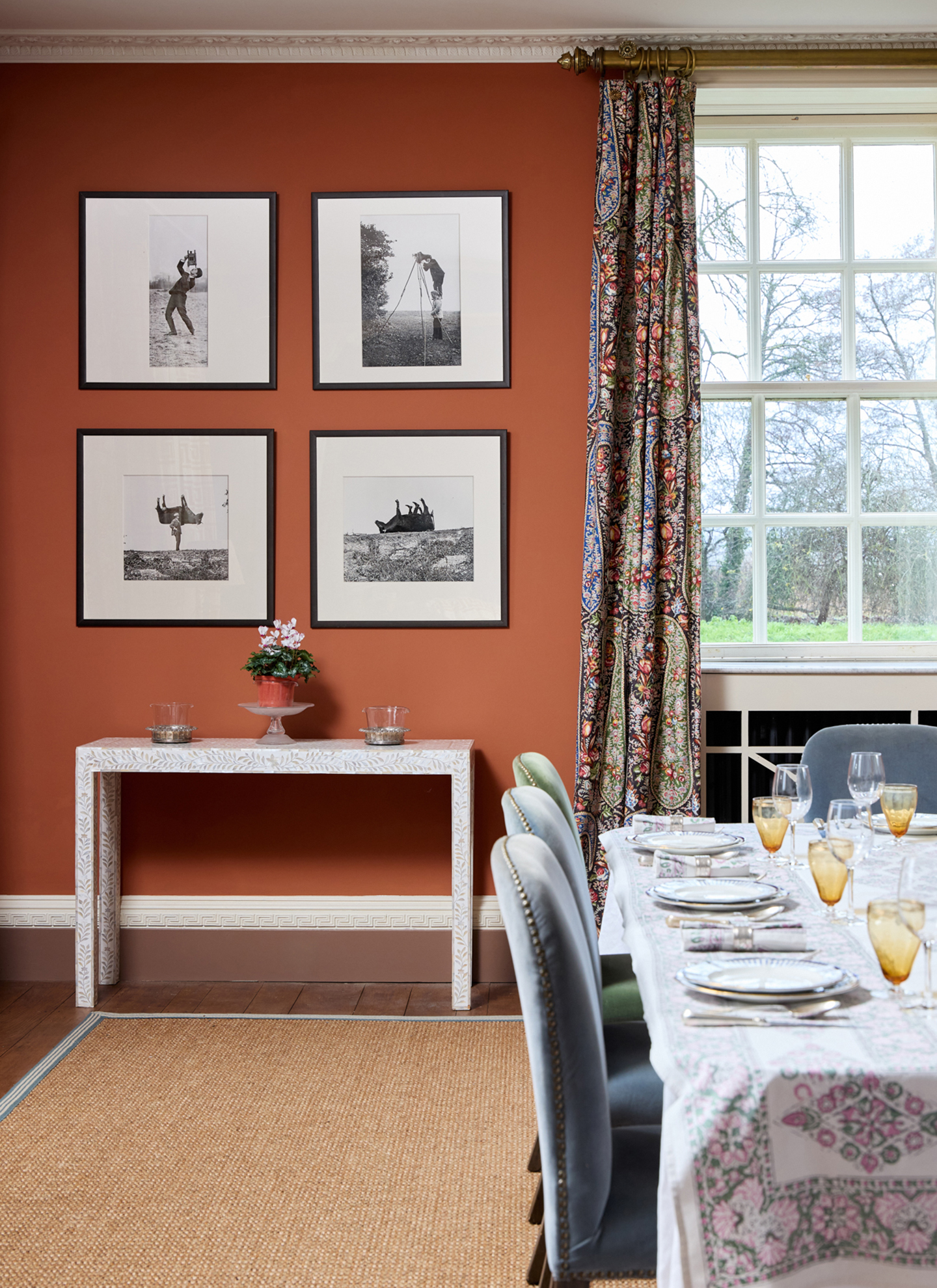 sophisticated dining room with terracotta orange walls, a grid of black-and-white framed prints, a white bone inlay console table, patterned curtains, a woven sisal rug, and a fully set table with plush velvet chairs