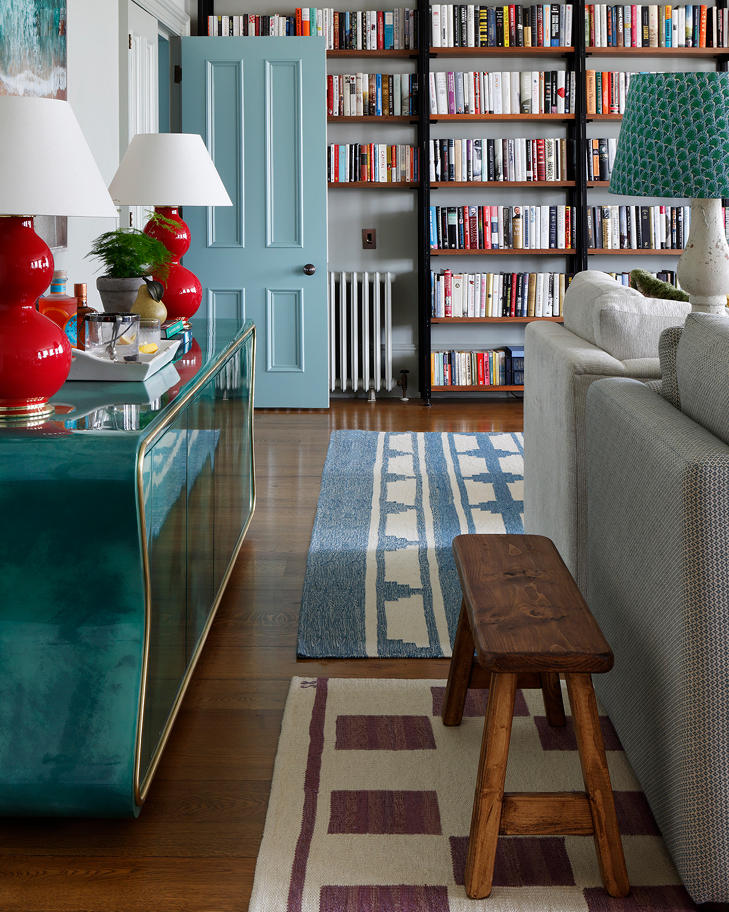 a living room with a large bookcase filled with books on the back wall, a shiny teal console table to the left with two large red shiny table lamps, a small wooden bench to the right and the backs of two grey sofas behind it