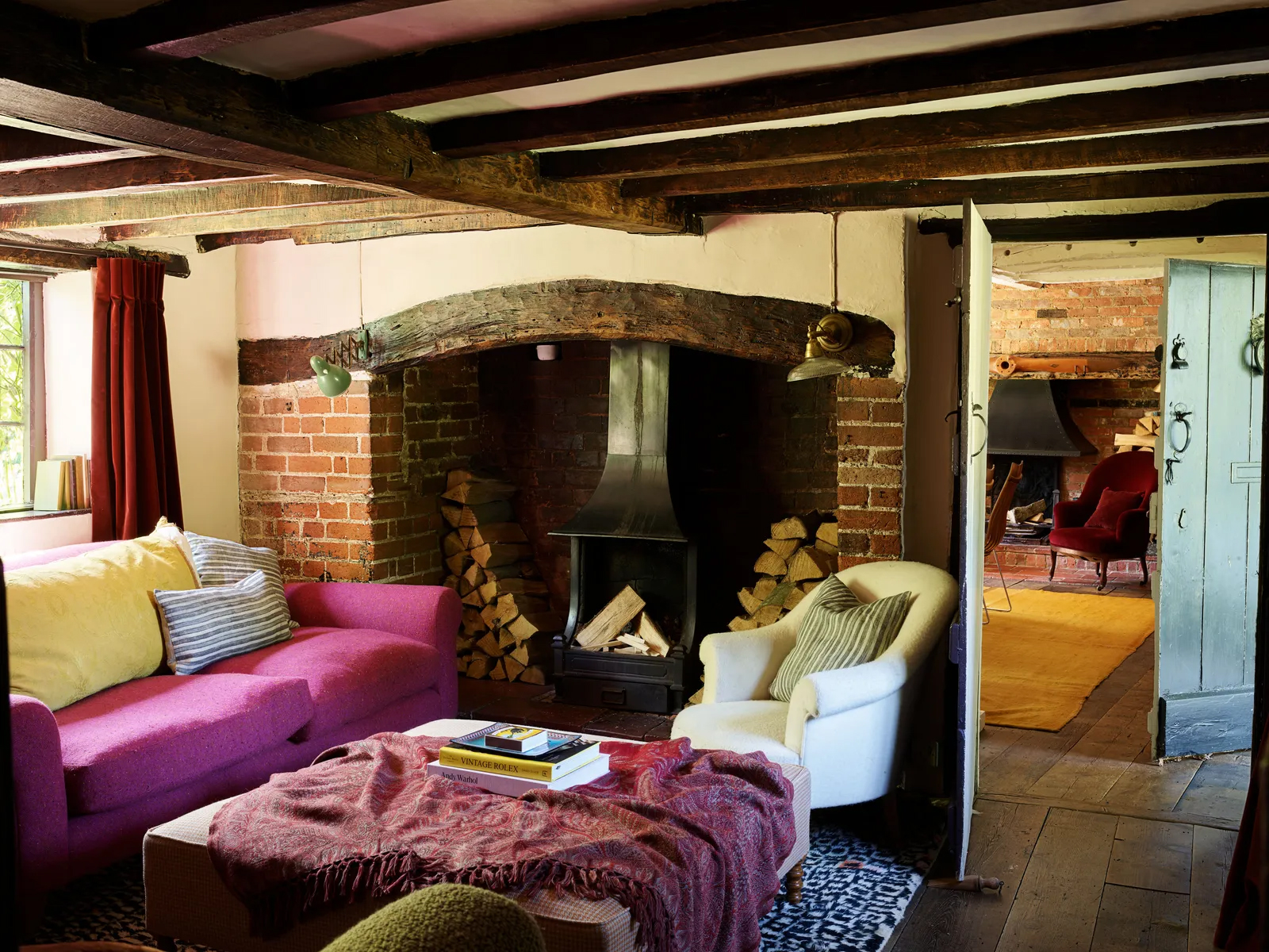 a cottage living room with wooden beams and red brick walls, a large fireplace, a pink sofa, a white armchair and an ottoman