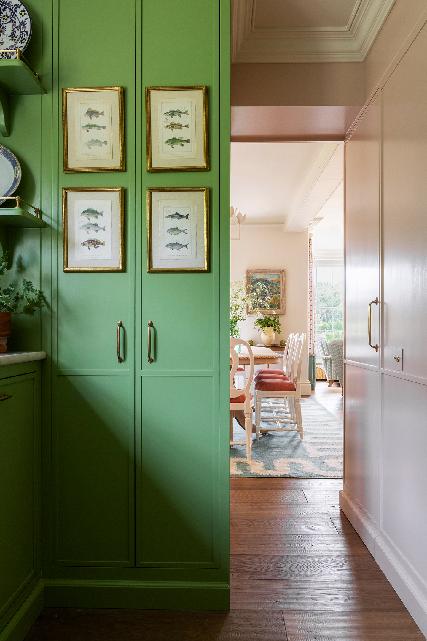 A pantry with tall green cupboards finished with brass handles and fish prints on the front doors. Down a hall to the right a dining area can be seen.