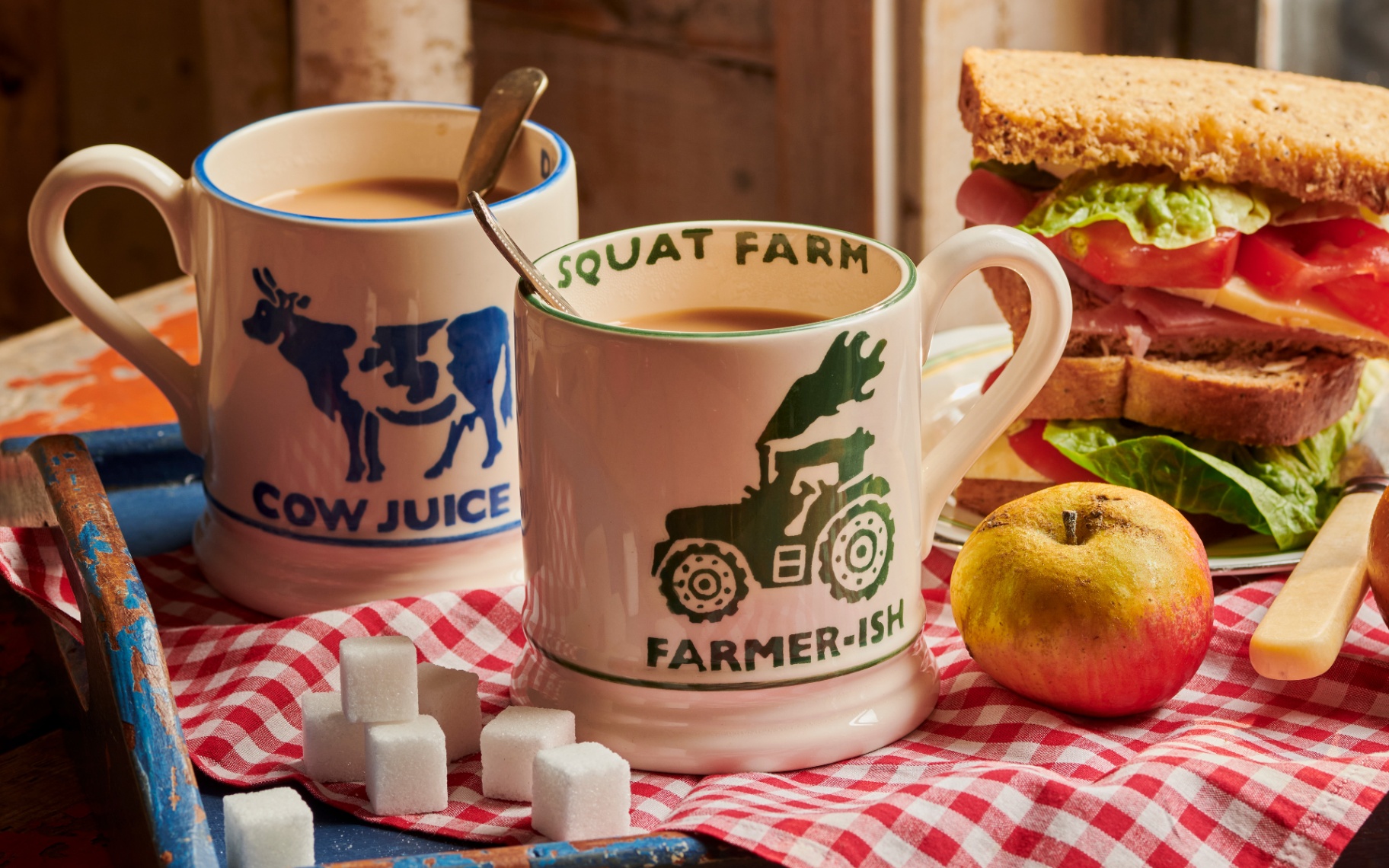 emma bridgewater diddly squat cow juice and farmer-ish mugs on a red gingham tea towel with a sandwich and an apple