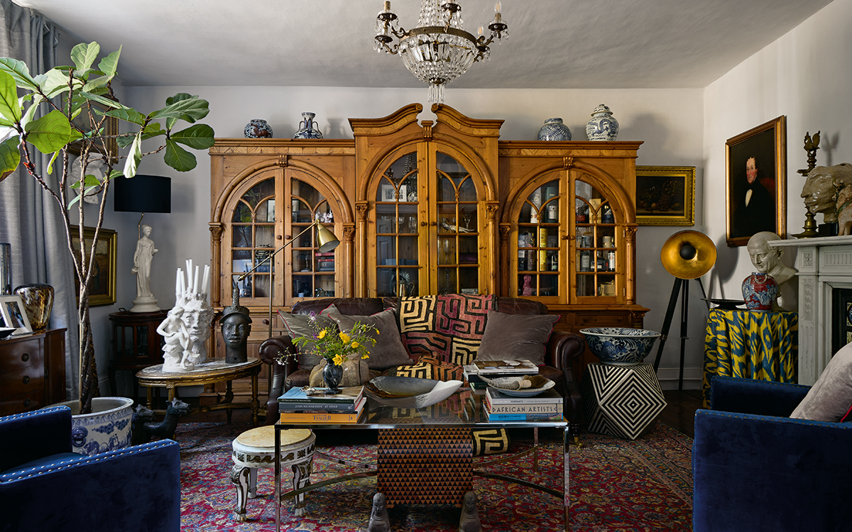living room filled with antique finds including a brown leather sofa dressed with cushions and a graphic throw, blue velvet armchairs, a red patterned rug, and extra large wooden bureau, above hangs a chandelier and to the left is a tall houseplant