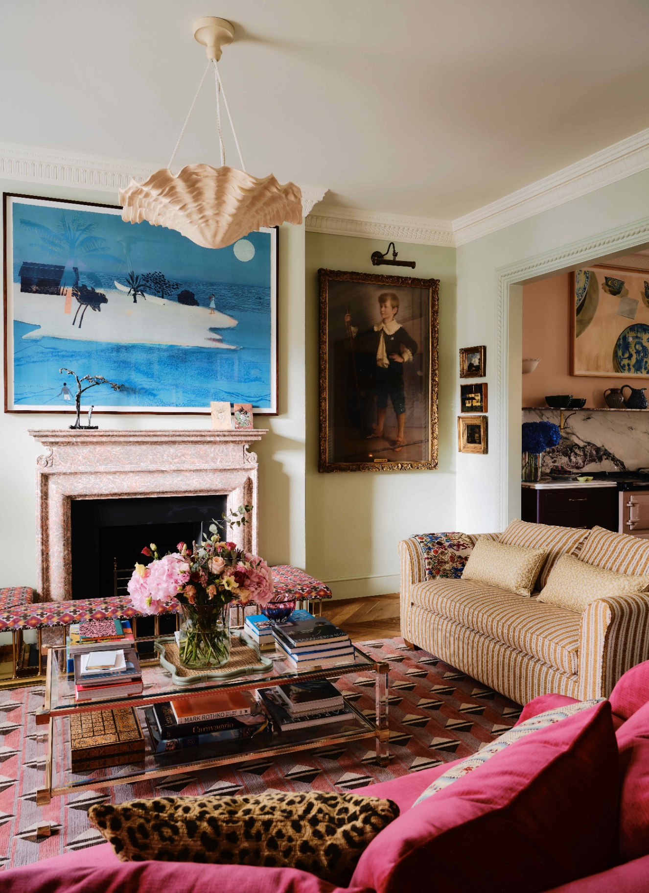 a living room with a marble fireplace with a large blue painting above it, a large wavy pendant light, a glass coffee table with stacks of coffee table books, a red sofa and a white and rust stripe sofa and a large portrait painting in a gold frame