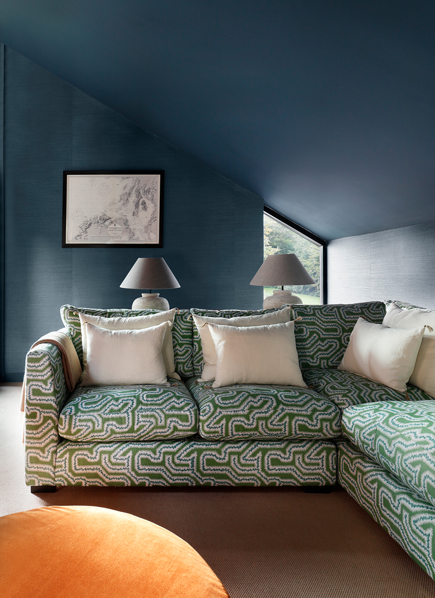 Dark blue attic room styled with a bold green patterned sectional sofa and white cushions. An orange velvet ottoman adds contrast in the foreground, with two matching lamps and framed map art against blue textured walls