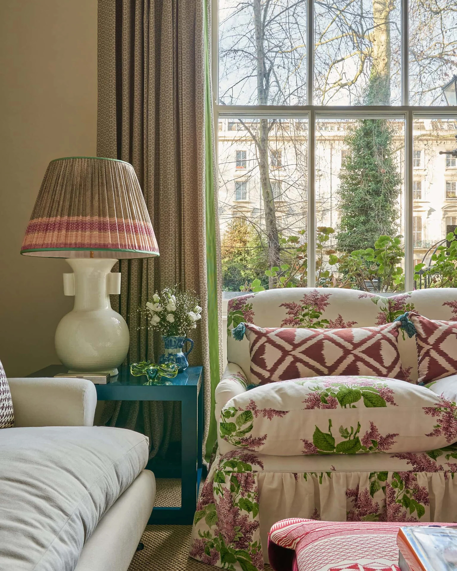 a living room corner with a large open window, a cream and pink floral skirted chair and a large pleated lamp on a side table