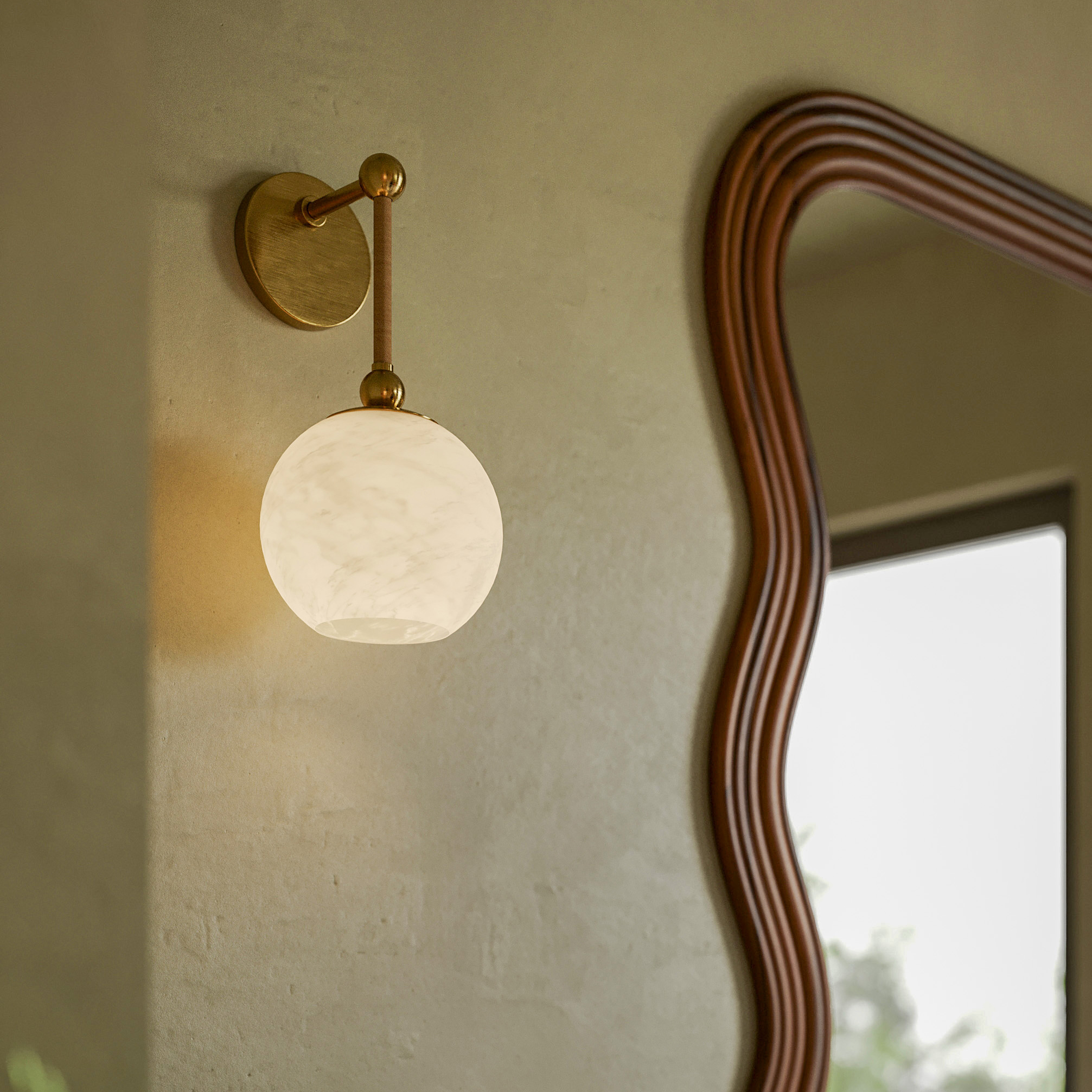 a gold wall sconce with a white glass globe shade on a green wall next to a wooden wavy edge mirror
