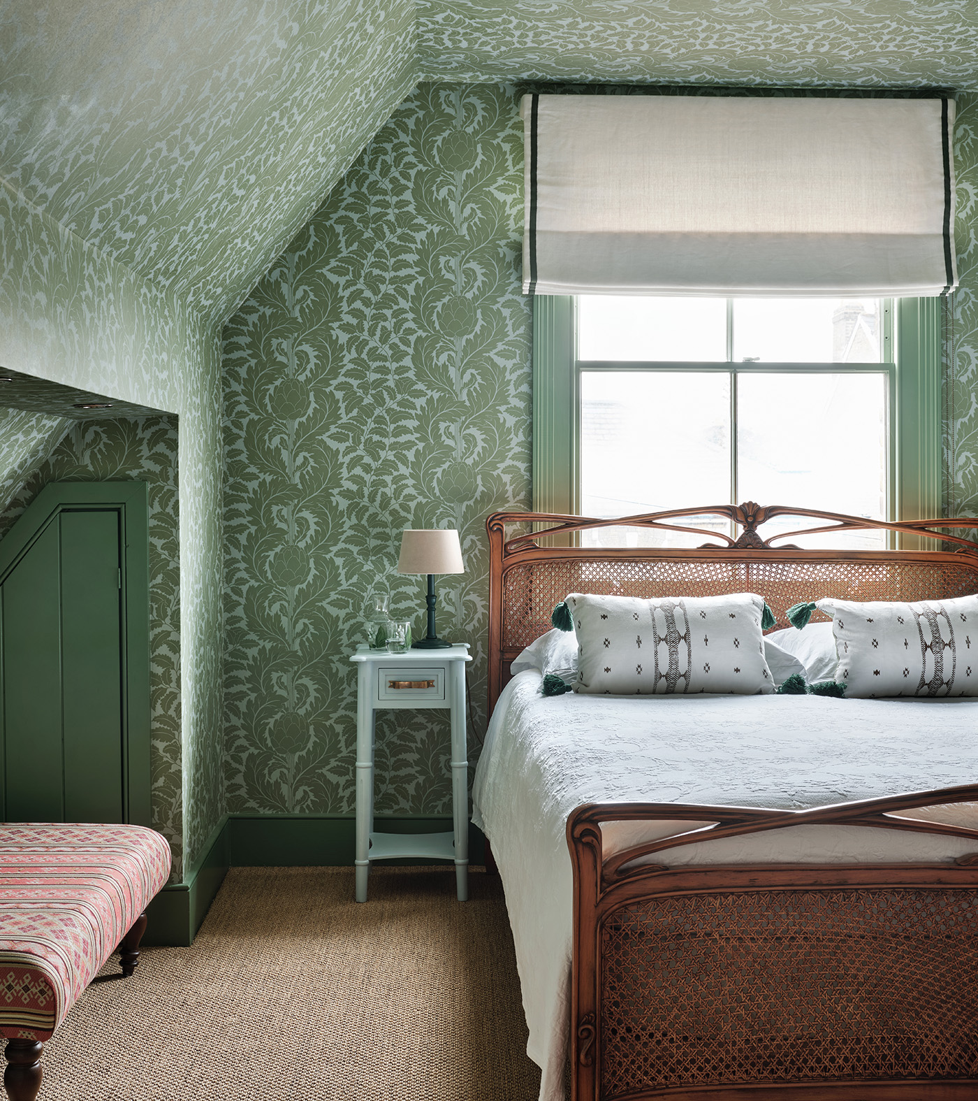 a bedroom with sloped ceilings, green botanical wallpaper on each wall and a rattan double bed with white bed linen