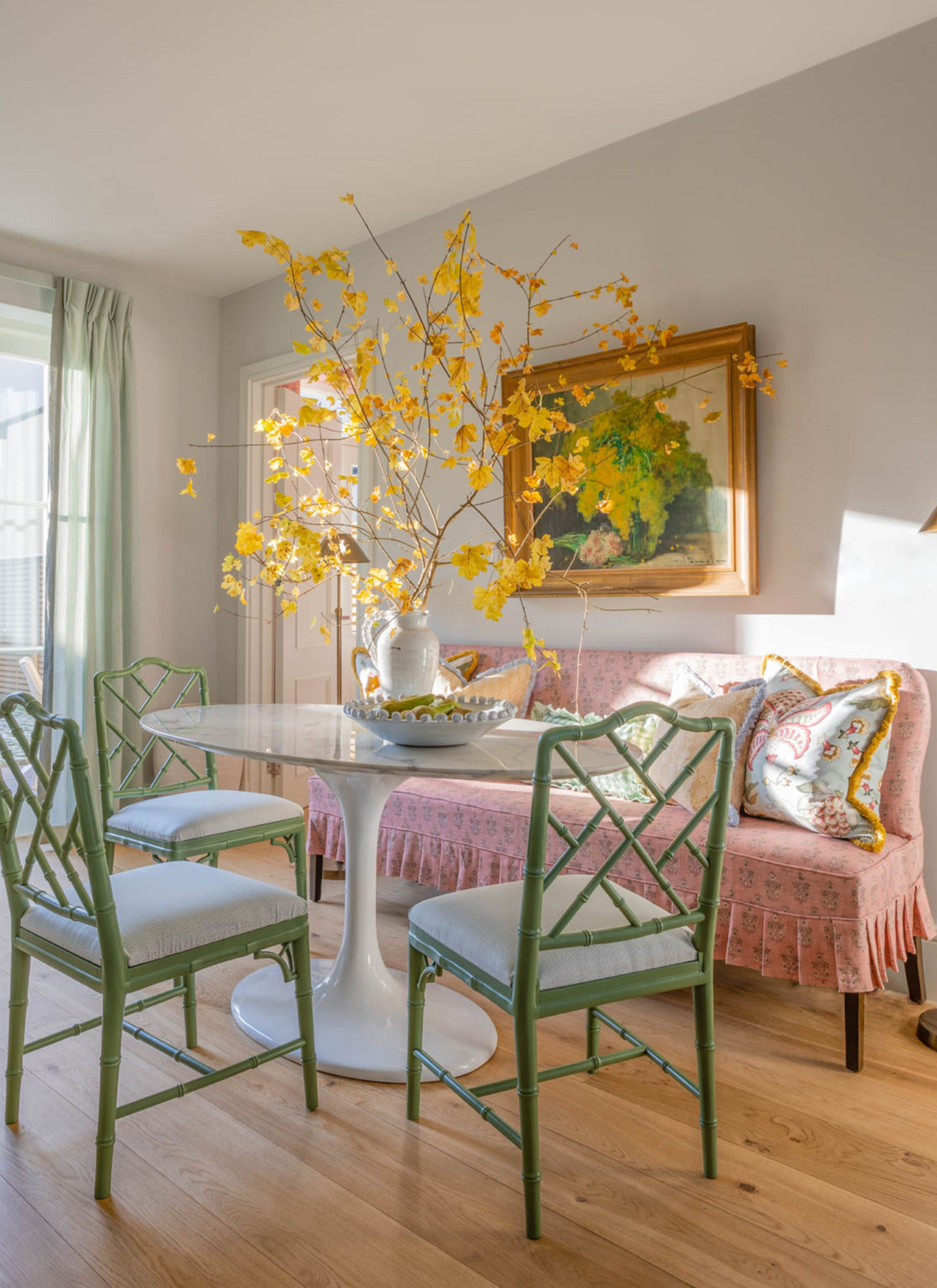 A marble-top dining table with a tall arrangement of sweeping, golden-leaved branches in a white ceramic pitcher, in a brightly lit room with a pink sofa and green chairs