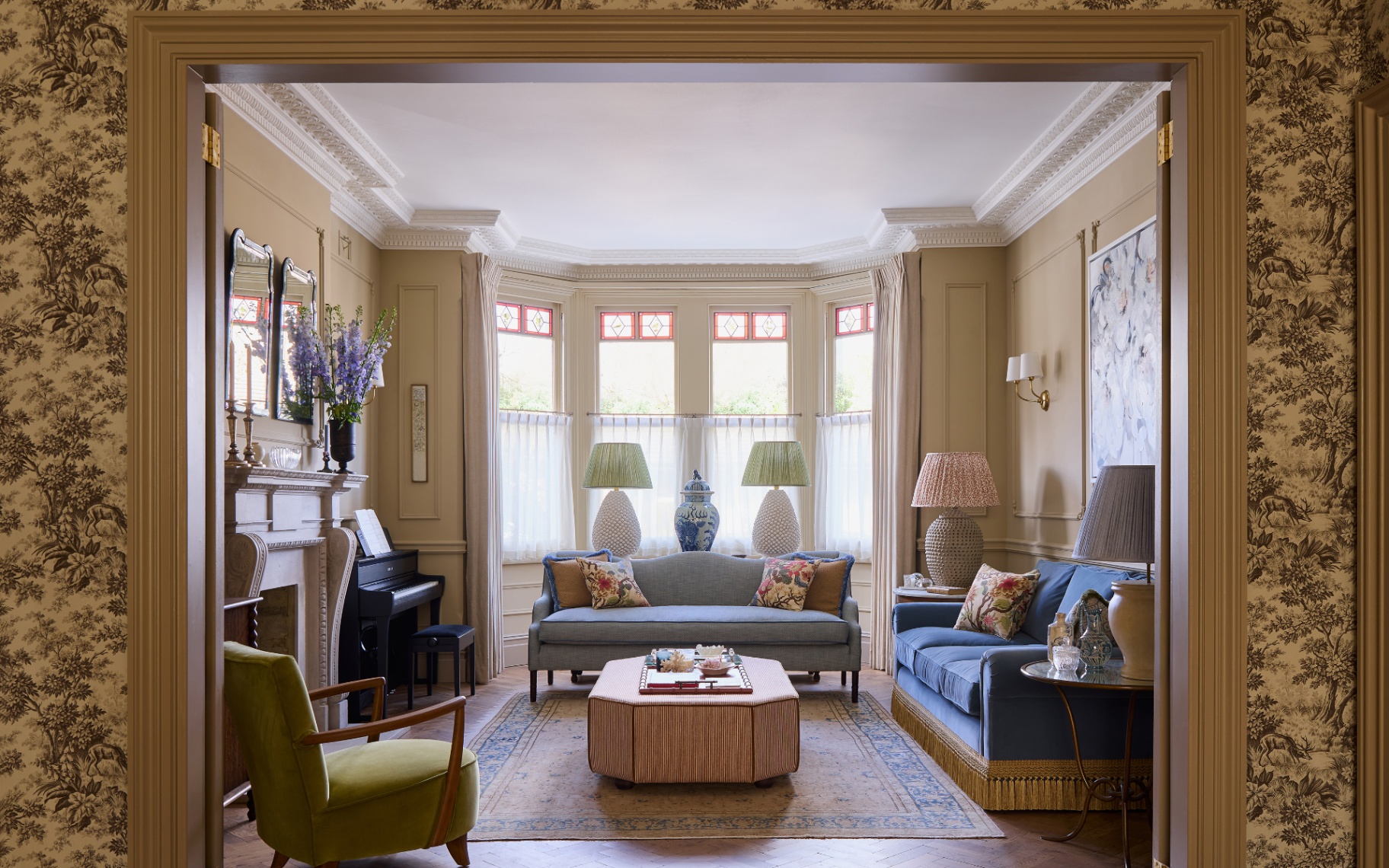 a living room with a wall with floral wallpaper, a large bay window at the back with a blue sofa in front of it, a fireplace and a piano to the left and a navy blue velvet sofa with yellow tassells to the right