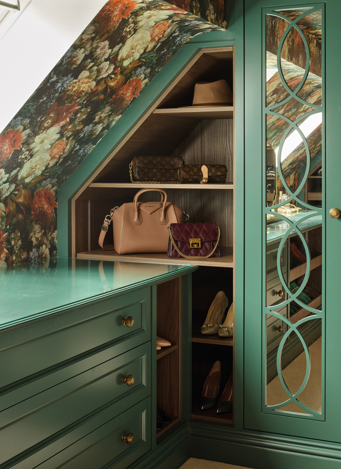a corner of a bedroom with green painted wardrobe and drawers, dark floral wallpaper and handbags on shelves