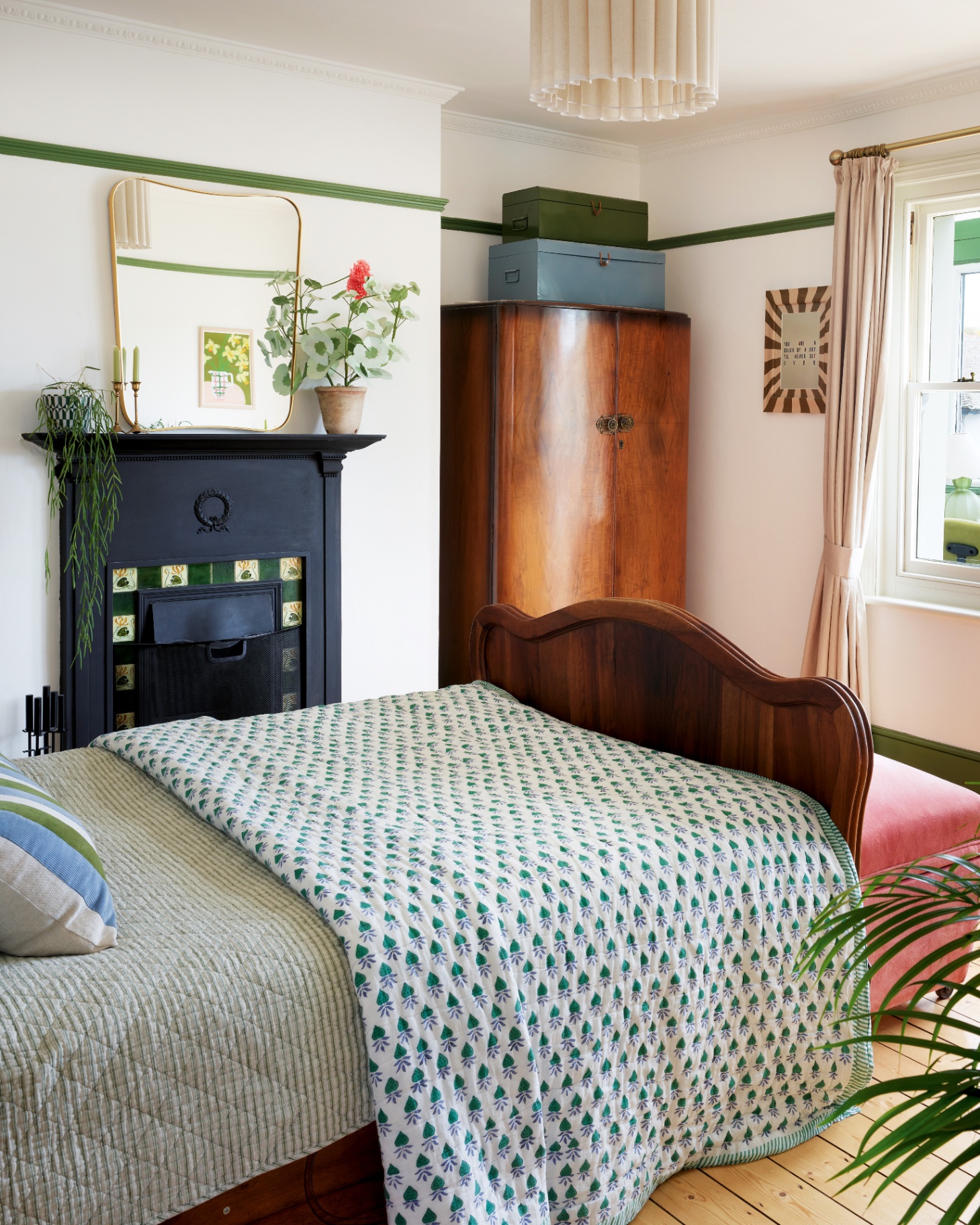 a bedroom with white walls and a mahogony wardrobe in the corner with two storage boxes on top, alarge black fireplace with a retro style mirror above it and a dark wood bed with a white blue and green patterned bedspread