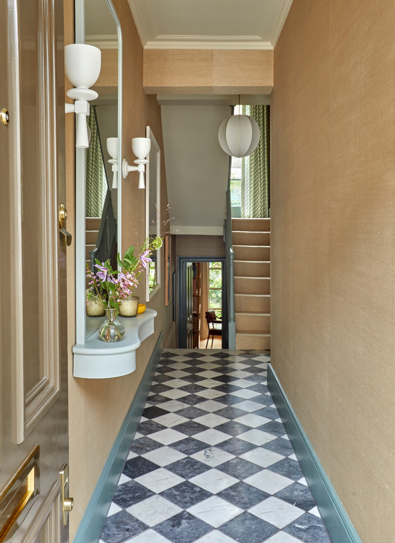 a hallway with beige walls, blakc and white checkerboard flooring and a large mirror on the left wall with a ledge with flowers and a candle