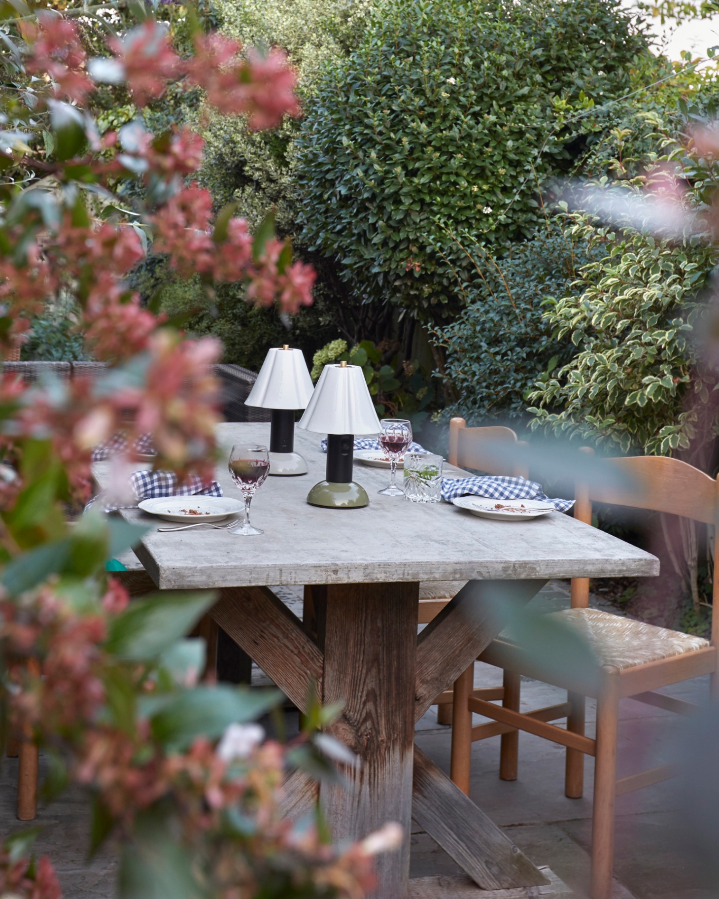 a large wooden outdoor table with apair of black an white rechargeable lamps on top, surrounded by green and red trees