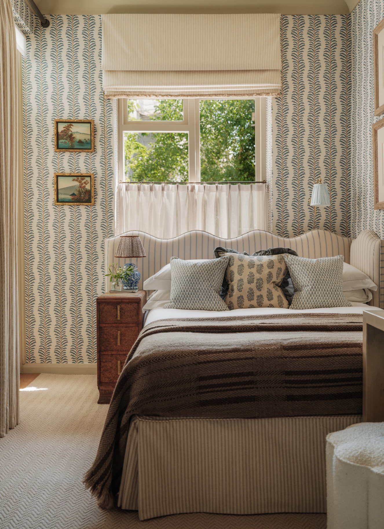 a bedroom with white and green leaf patterned wallpaper, a large bed with astriped upholstered headboard and valance and a window with a roman blind and a cafe curtain