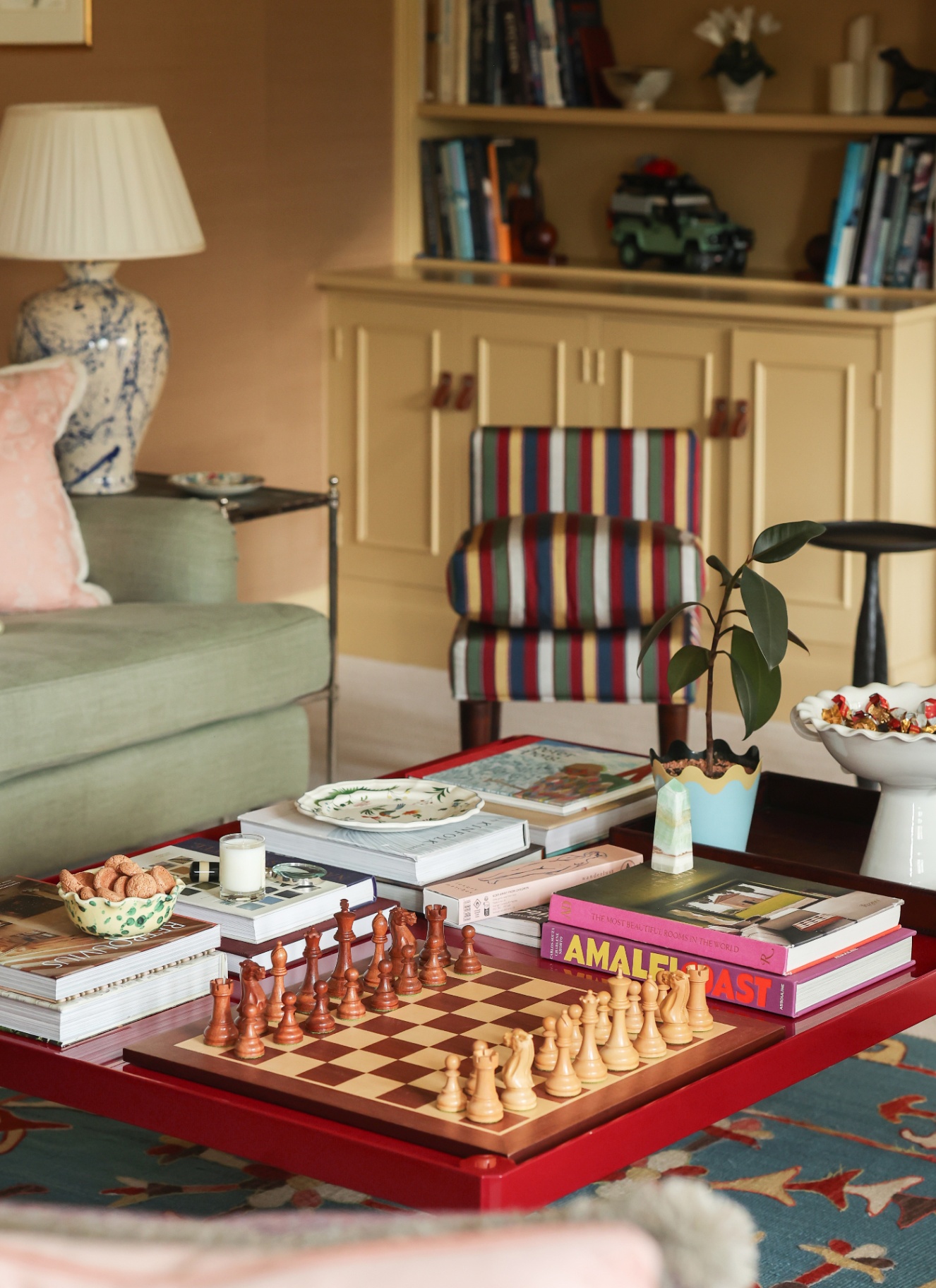 A colorful sitting room with a red coffee table setting showcasing a wooden chessboard, stack of art books, a striped accent chair, a pale green sofa, and cream built-in cabinetry.