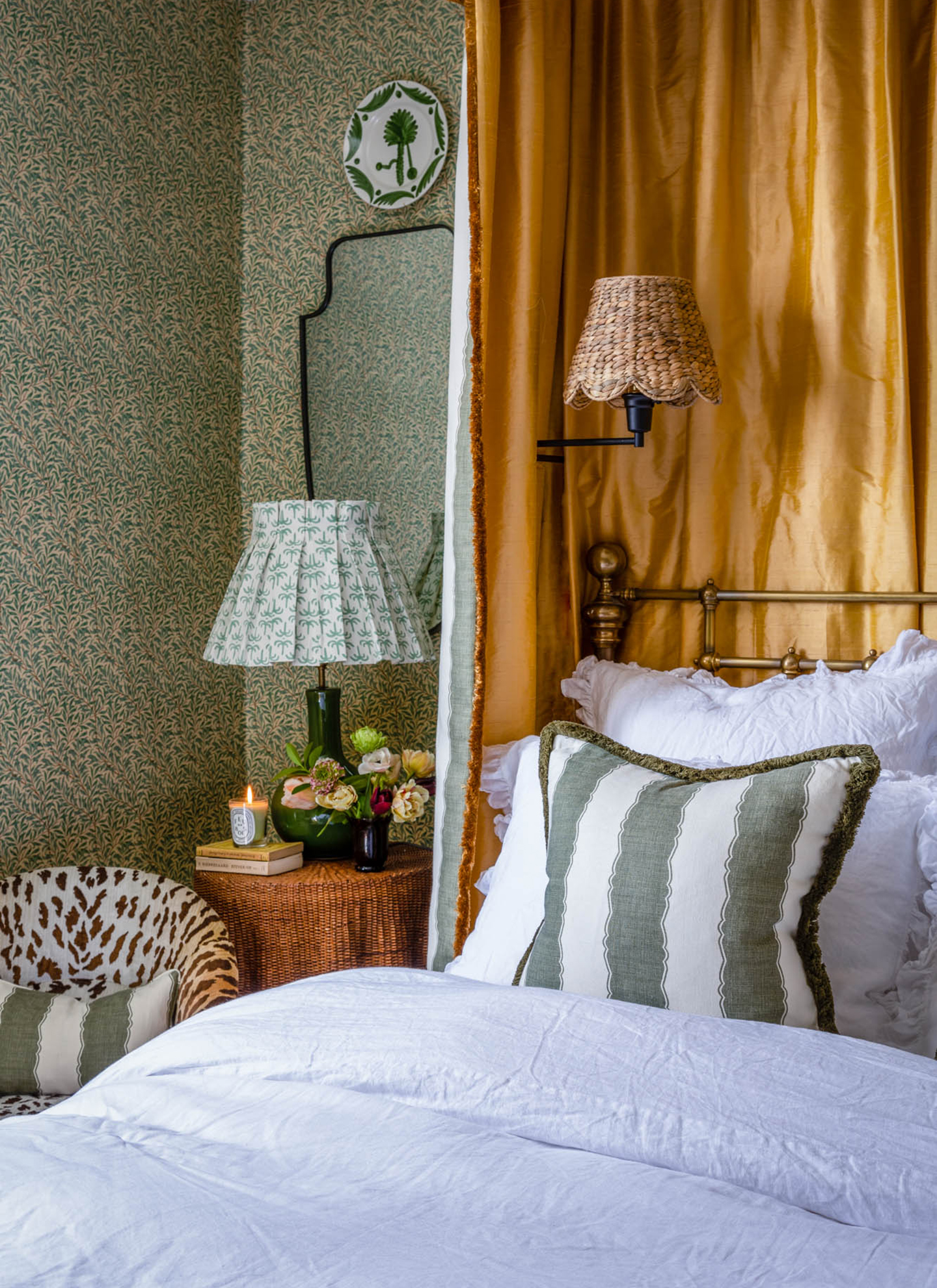 A bedside table with a green lamp, books, a burning candle, and a small posy of cream and pink blooms, next to a bed with gold canopy curtains and patterned wallpaper