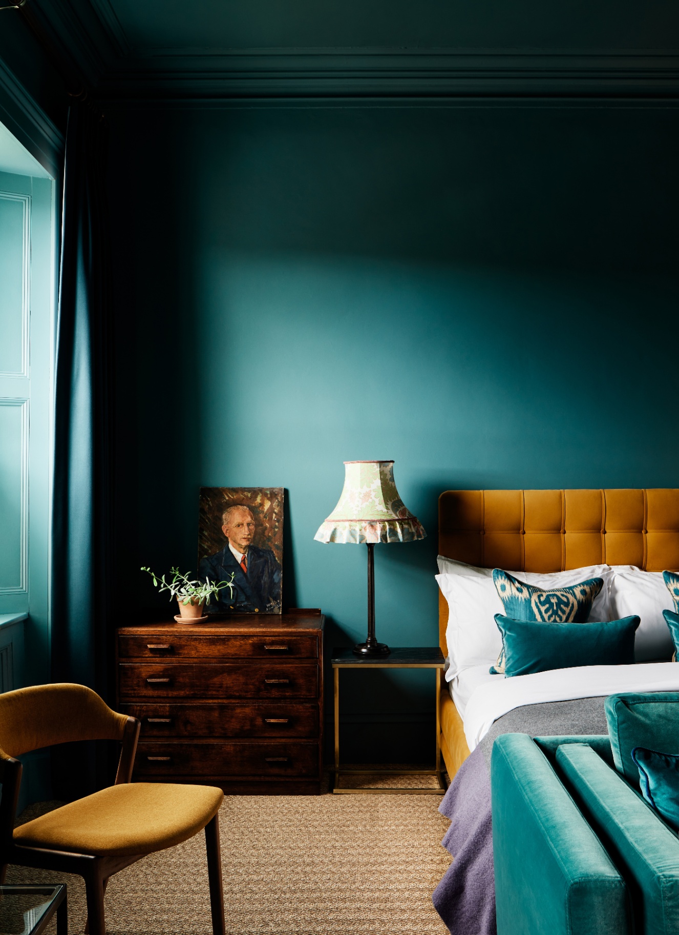 Deep petrol blue room with color-drenched walls and ceiling, contrasted by a bright mustard yellow tufted velvet headboard and a vintage wooden chest of drawers