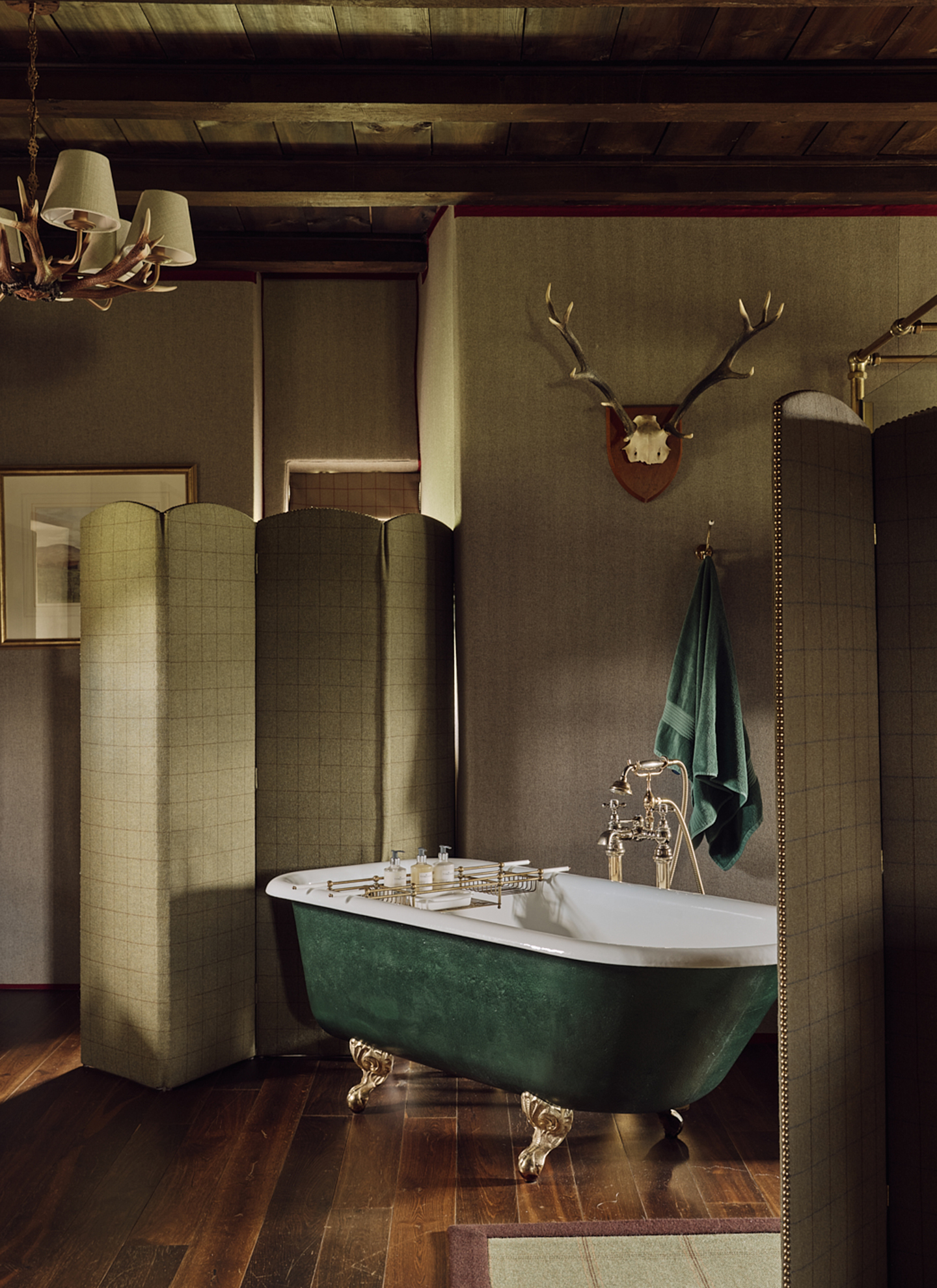 An atmospheric, rustic bathroom featuring a deep green clawfoot tub with brass fittings, set against textured olive walls, dark wood flooring, and a folding tweed room divider.