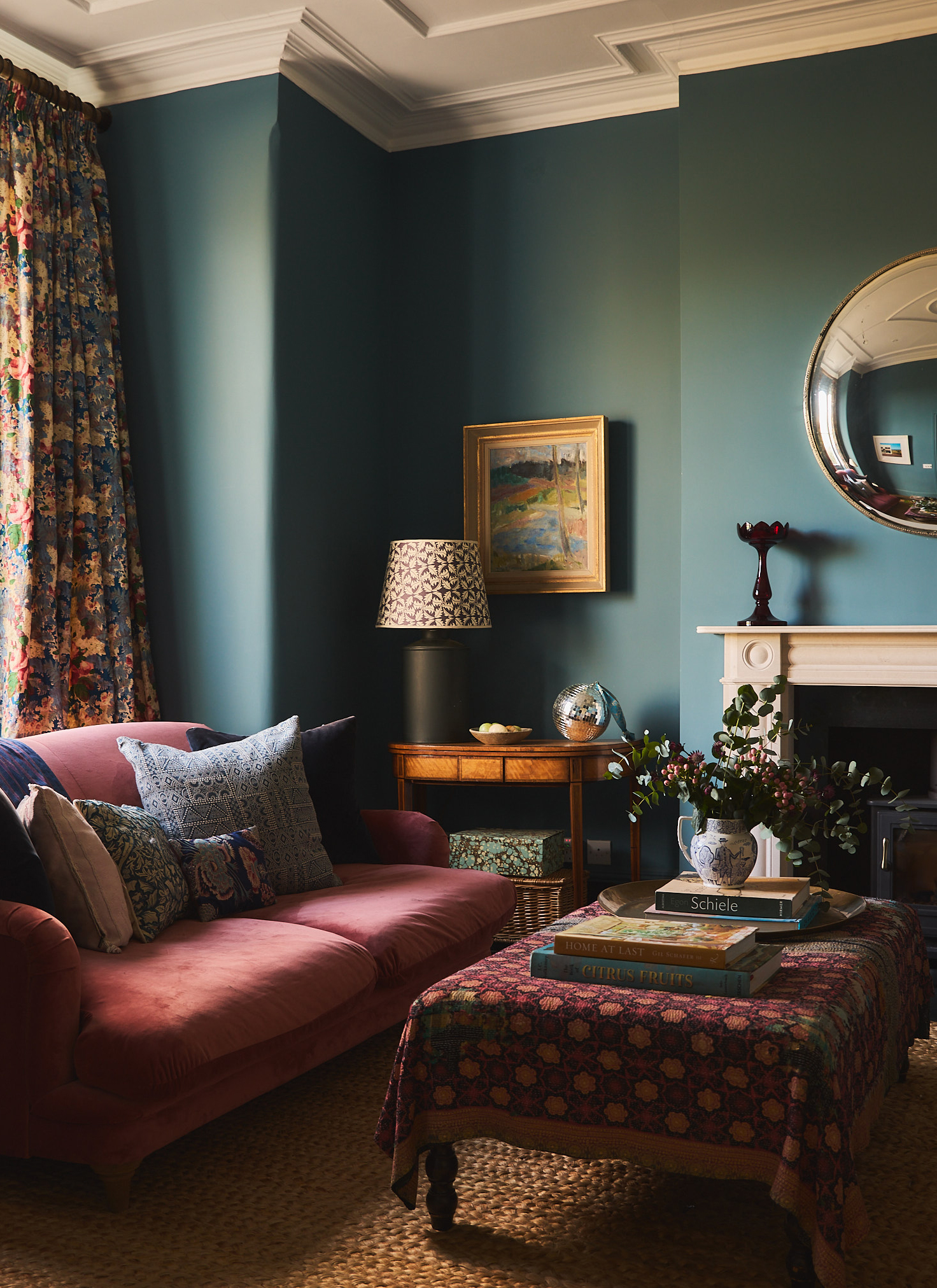 An elegant sitting room with deep teal walls, a pink velvet sofa adorned with patterned cushions, a patterned ottoman coffee table, and floral curtains beside a fireplace