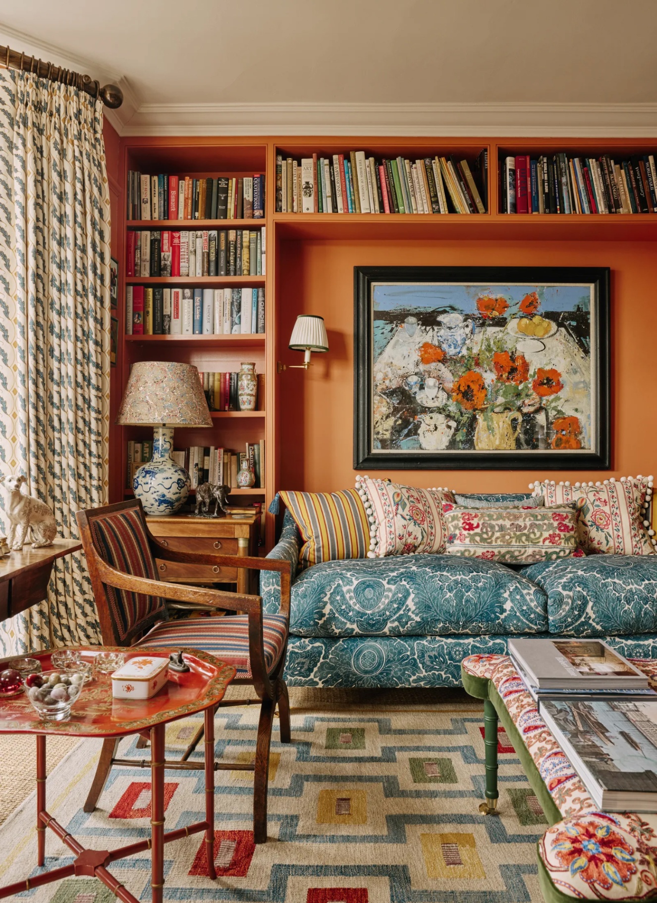 An eclectic reading corner featuring terracotta painted built-in bookshelves, a blue floral patterned sofa with patterned cushions, a geometric rug, and framed artwork