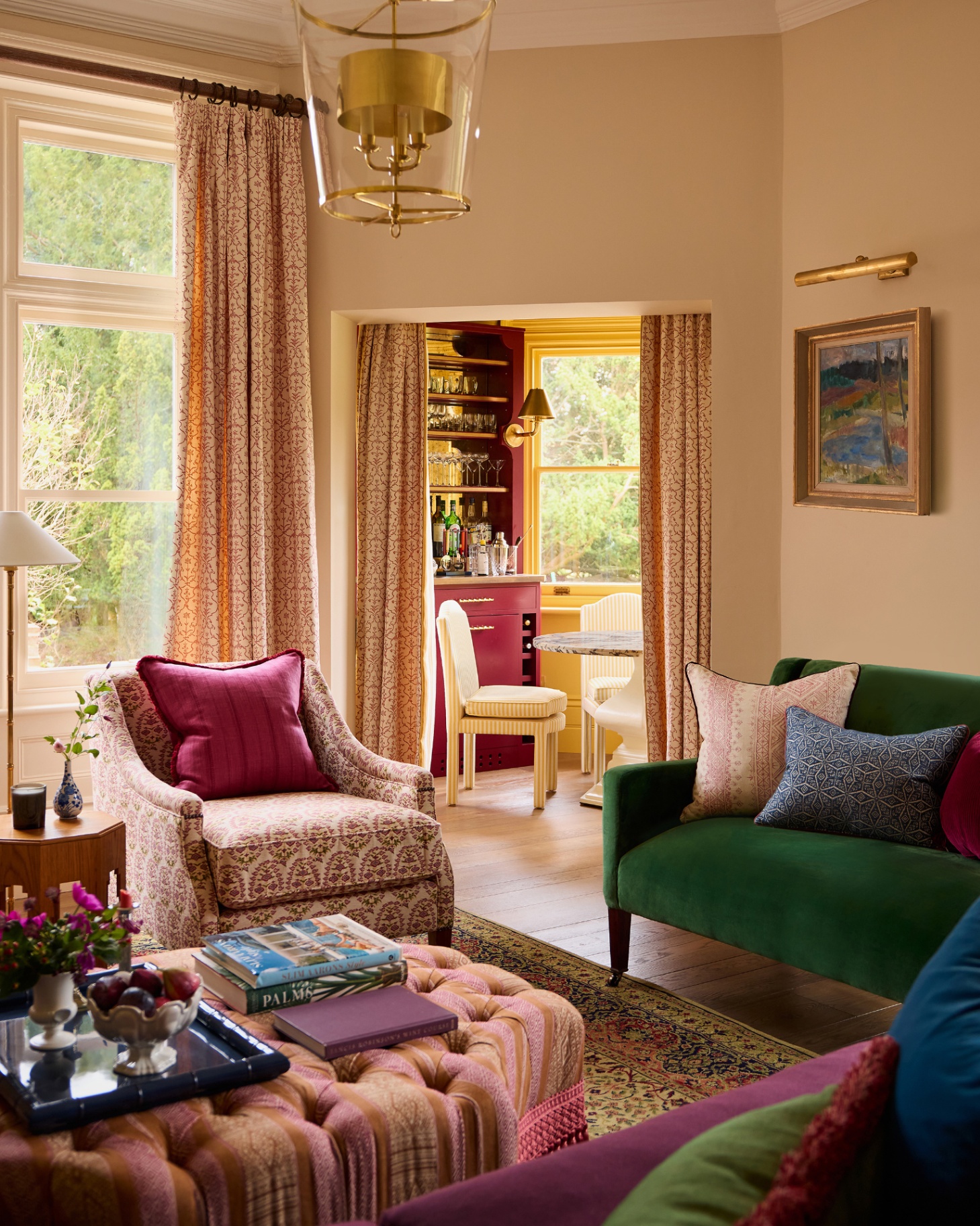 a living room with a emerald green sofa, a white and red patterned armchair, an ottoman and an open door to a dining room