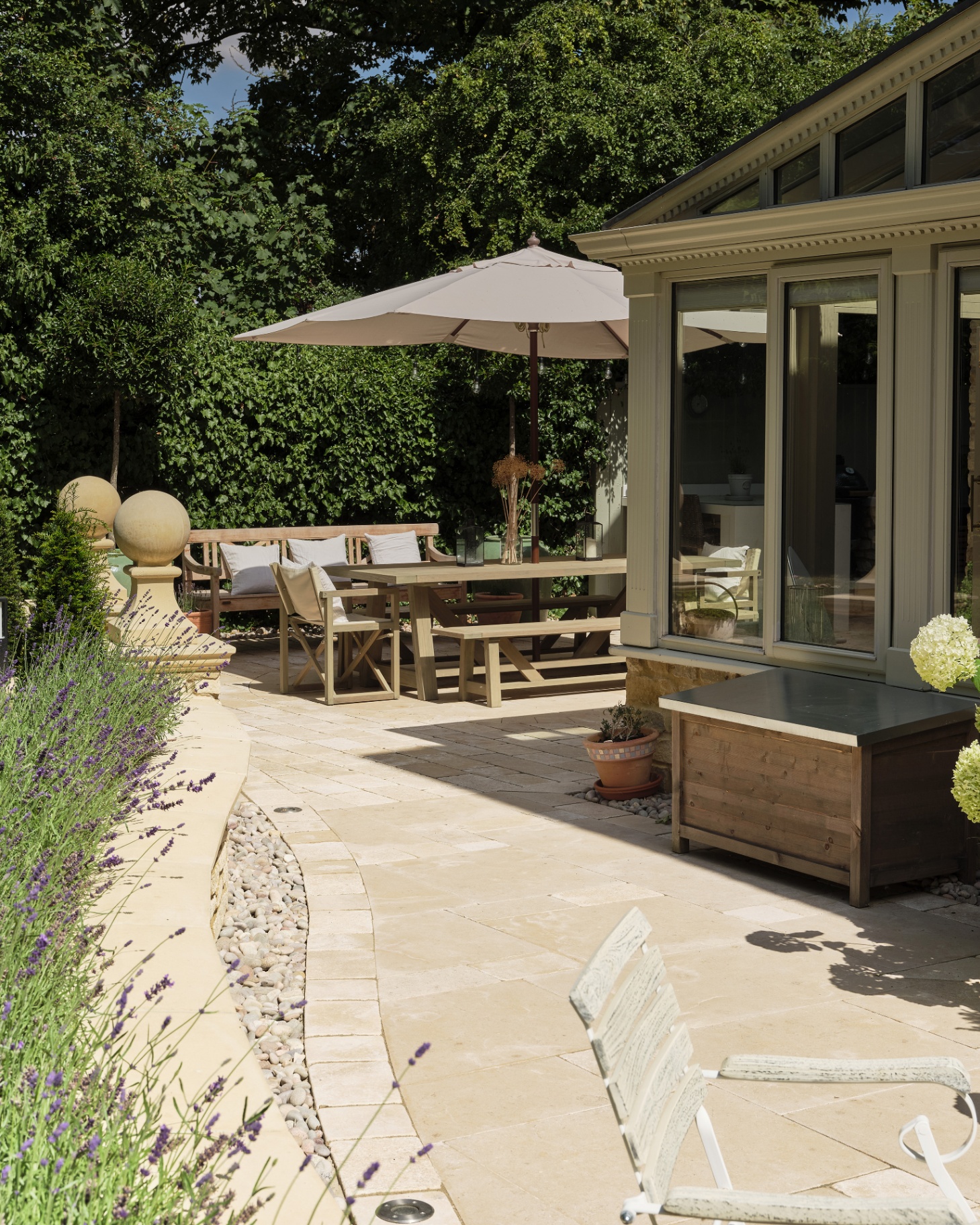 a garden with a beige stone patio, a green conservatory and an outdoor dining set with a cream parasol