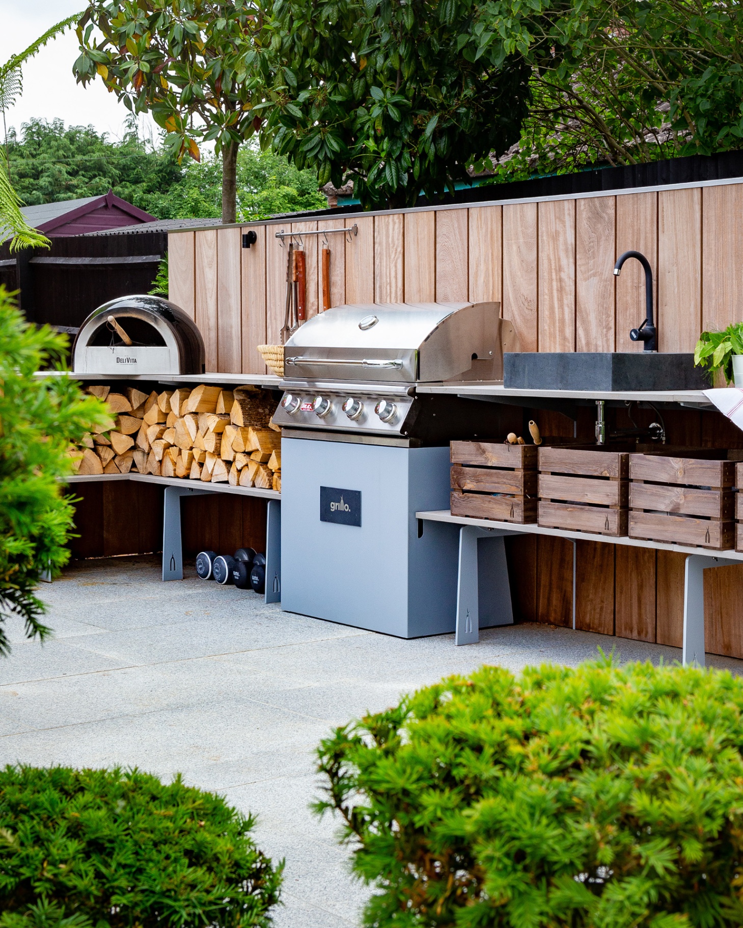 outdoor kitchen area with a pizza oven, a bbq, logs stacked under the worktop, a grey patio and trees behind the kitchen and in the foreground