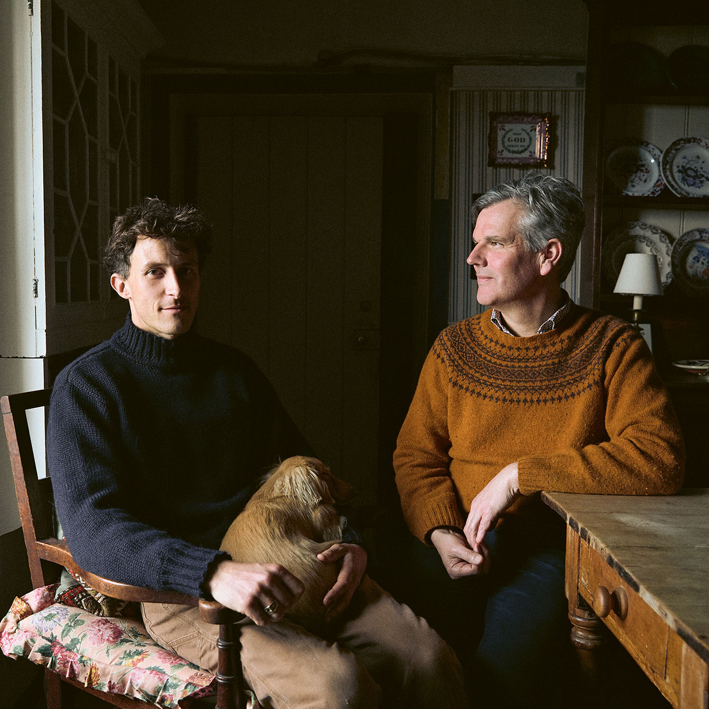 two men sit in a kitchen, one with a dog on his lap