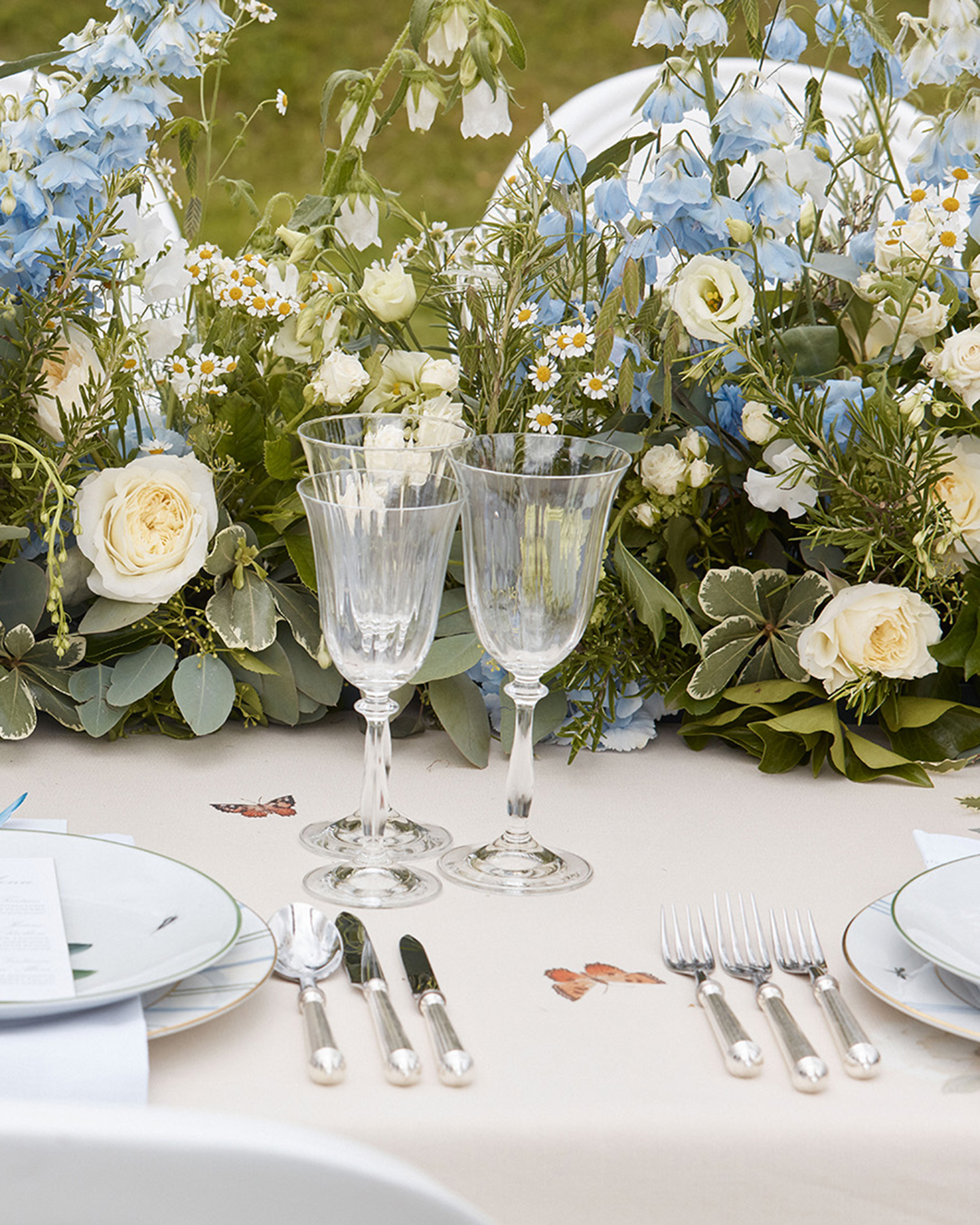 a summer tablescape with blue and white fresh flowers, clear crystal wine glasses, a butterfly patterned tablecloth and silver cutlery